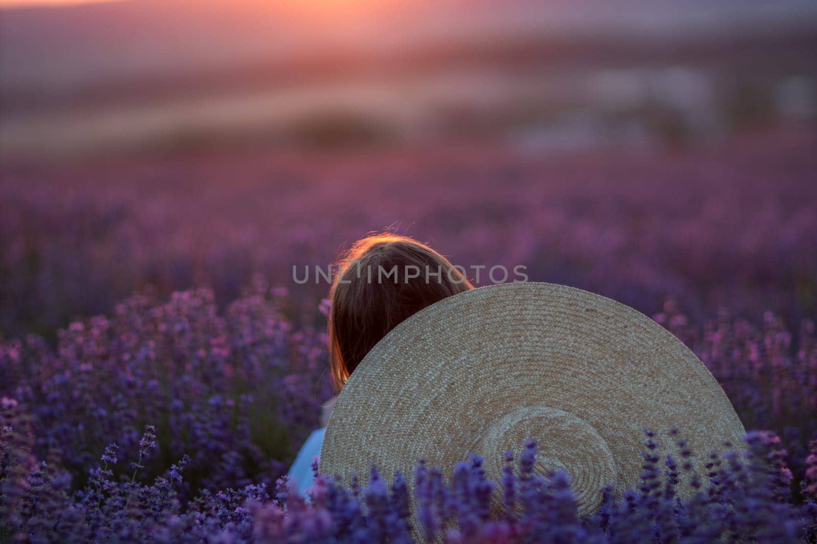 Here's a caption for the image: Woman with brown hair sits amidst a vast lavender field at sunset, a large straw hat resting on her back. The setting sun casts a warm glow.