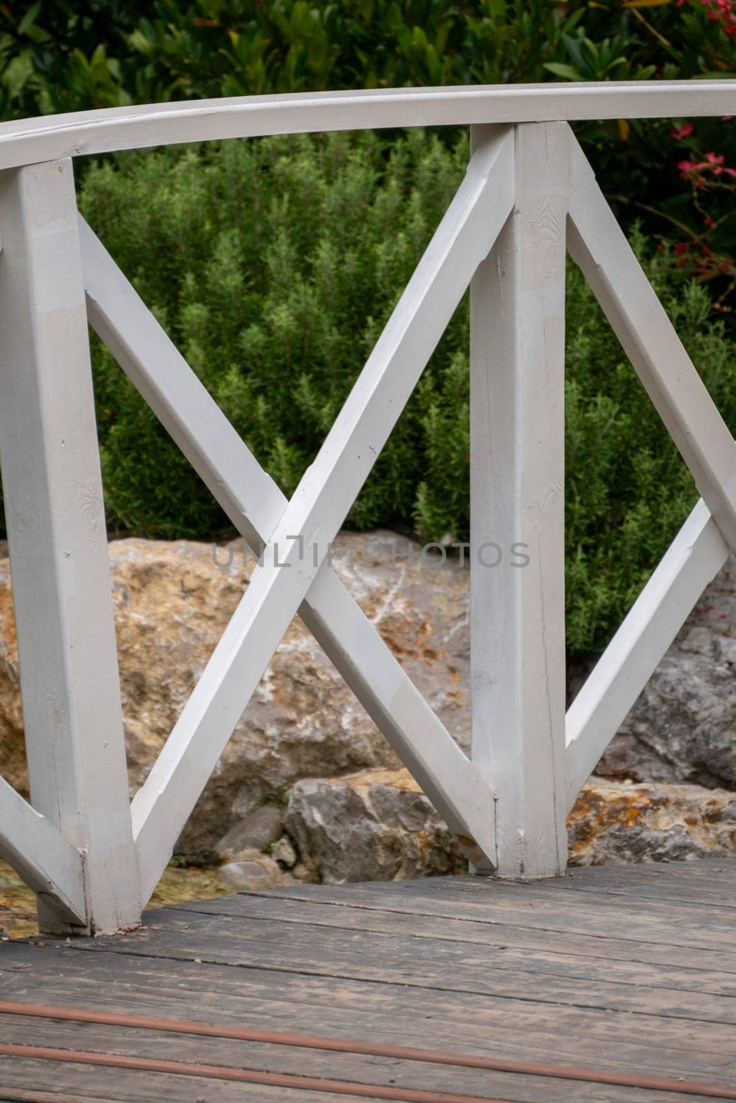 A close-up of a white wooden bridge railing with diamond-shaped panels. The railing is over a rocky area with green foliage in the background.