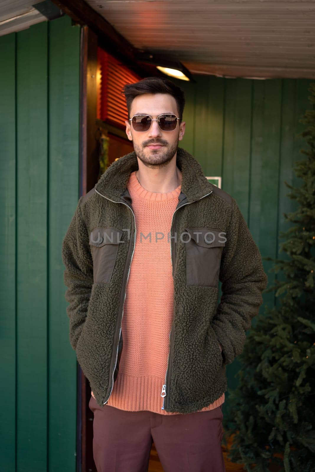 Confident handsome man in warm jacket standing with hands in pockets outside green house. Young casual good looking male is posing outdoors during Christmas.