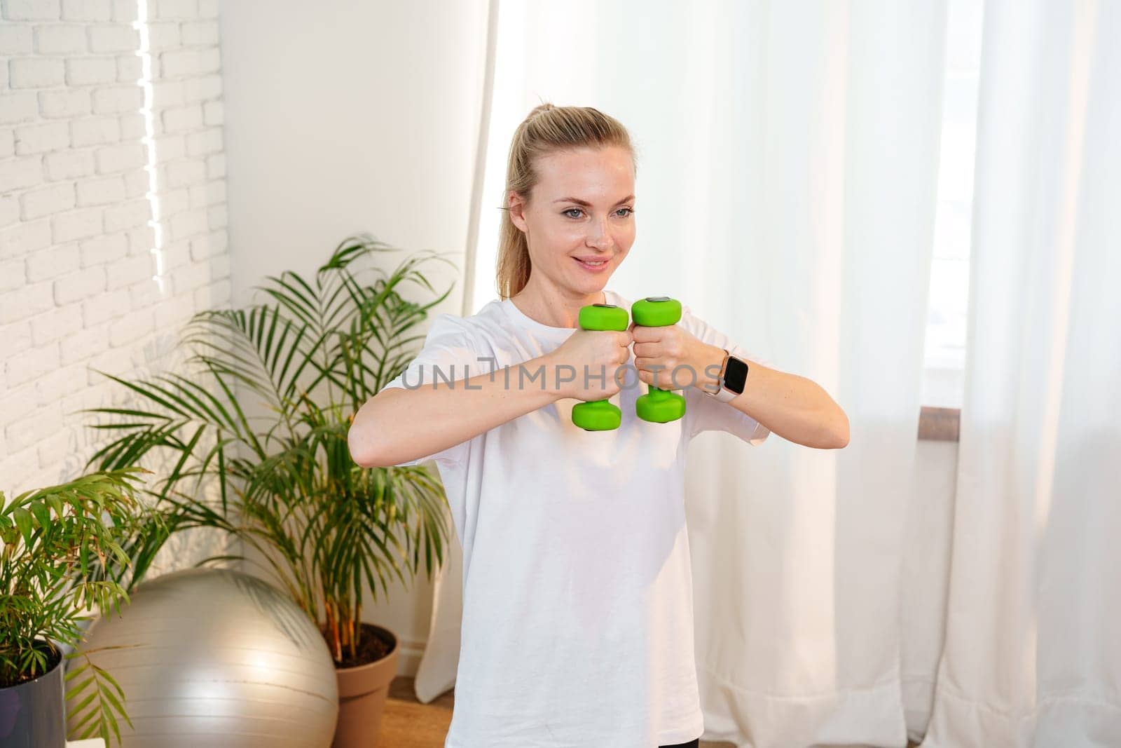 A woman exercises with green dumbbells at home in a bright room filled with light and plants