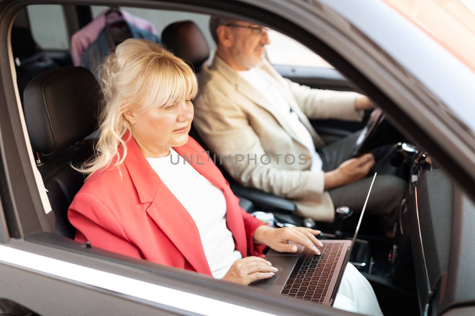 A woman in a red blazer works on her laptop while sitting in a car. A man drives, showcasing a blend of business and travel in a busy urban environment.
