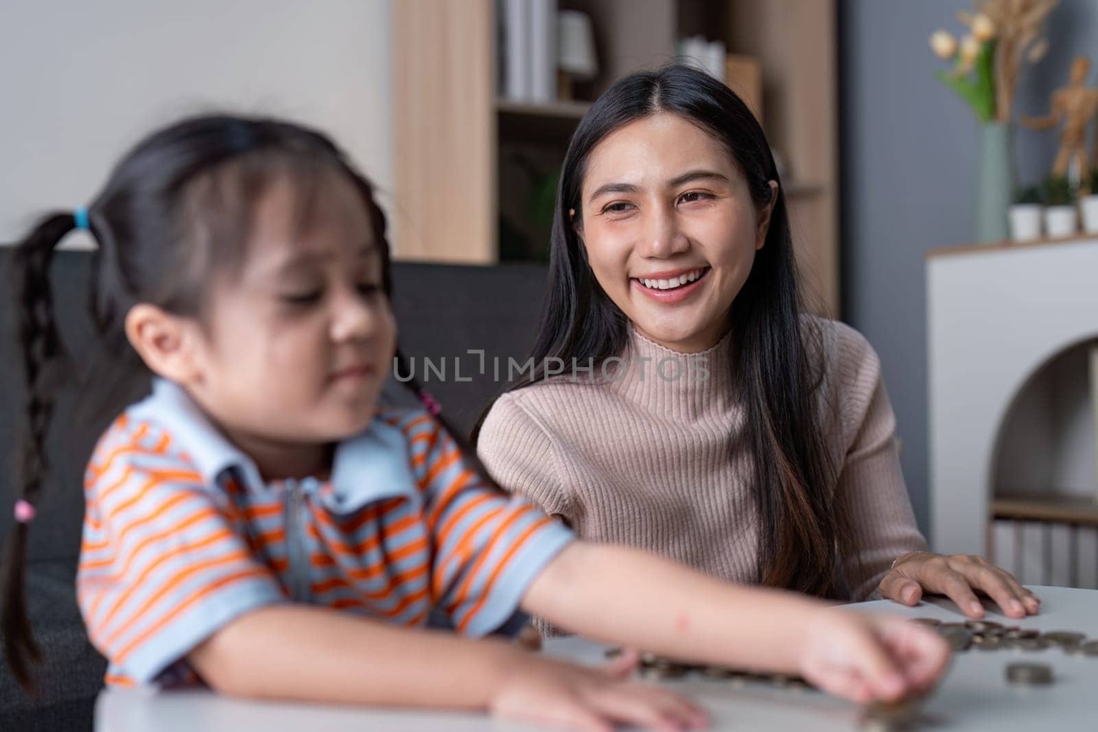 Asian mother smiles proudly watching her daughter sort coins, encouraging financial education and bonding at home with a pink piggy bank