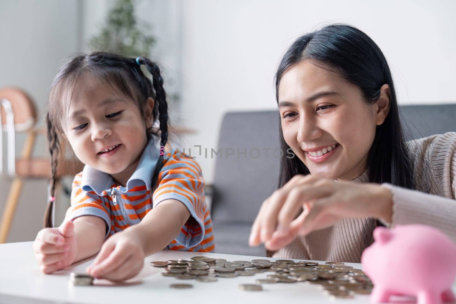 Asian mother and daughter happily sort coins together, fostering a loving financial learning experience at home using a pink piggy bank.