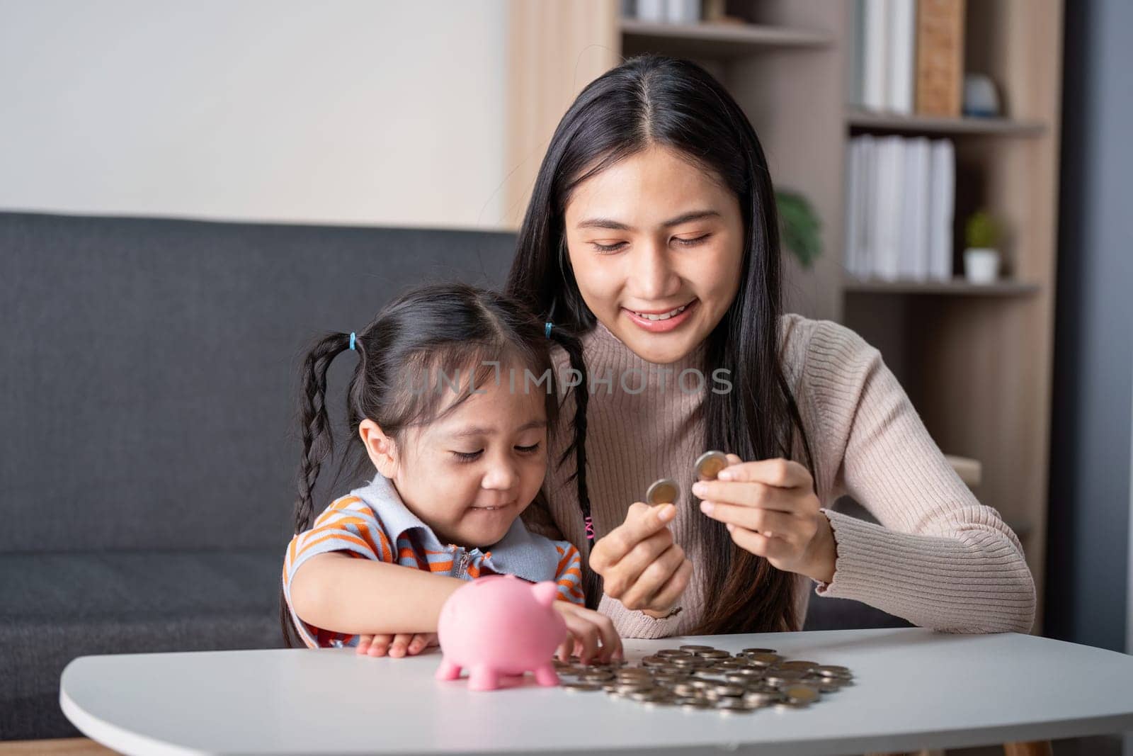 Asian mother and daughter share a bonding moment counting coins and saving them in a pink piggy bank, enhancing learning at home.