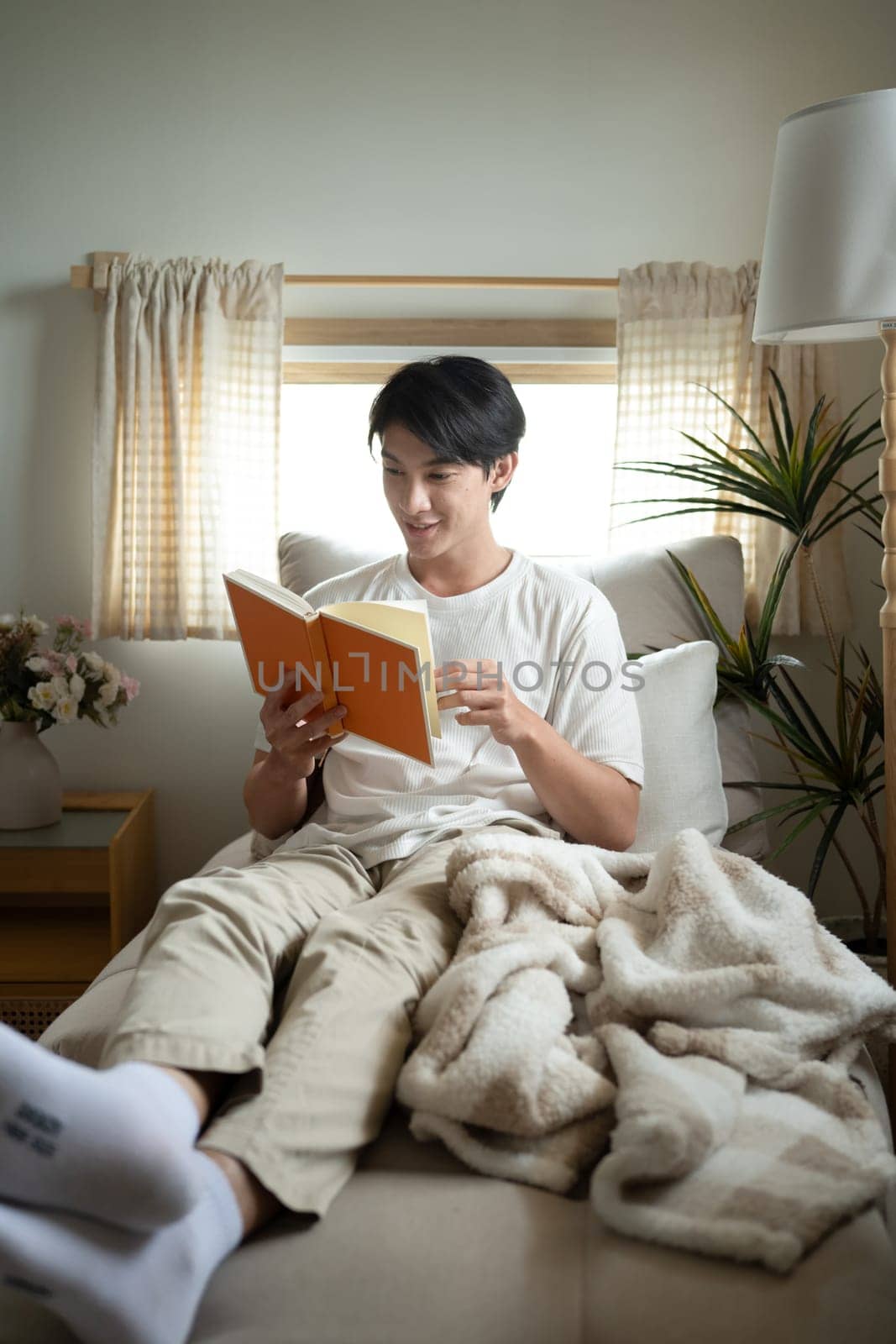 Peaceful millennial man reading book while relaxing on sofa at home.