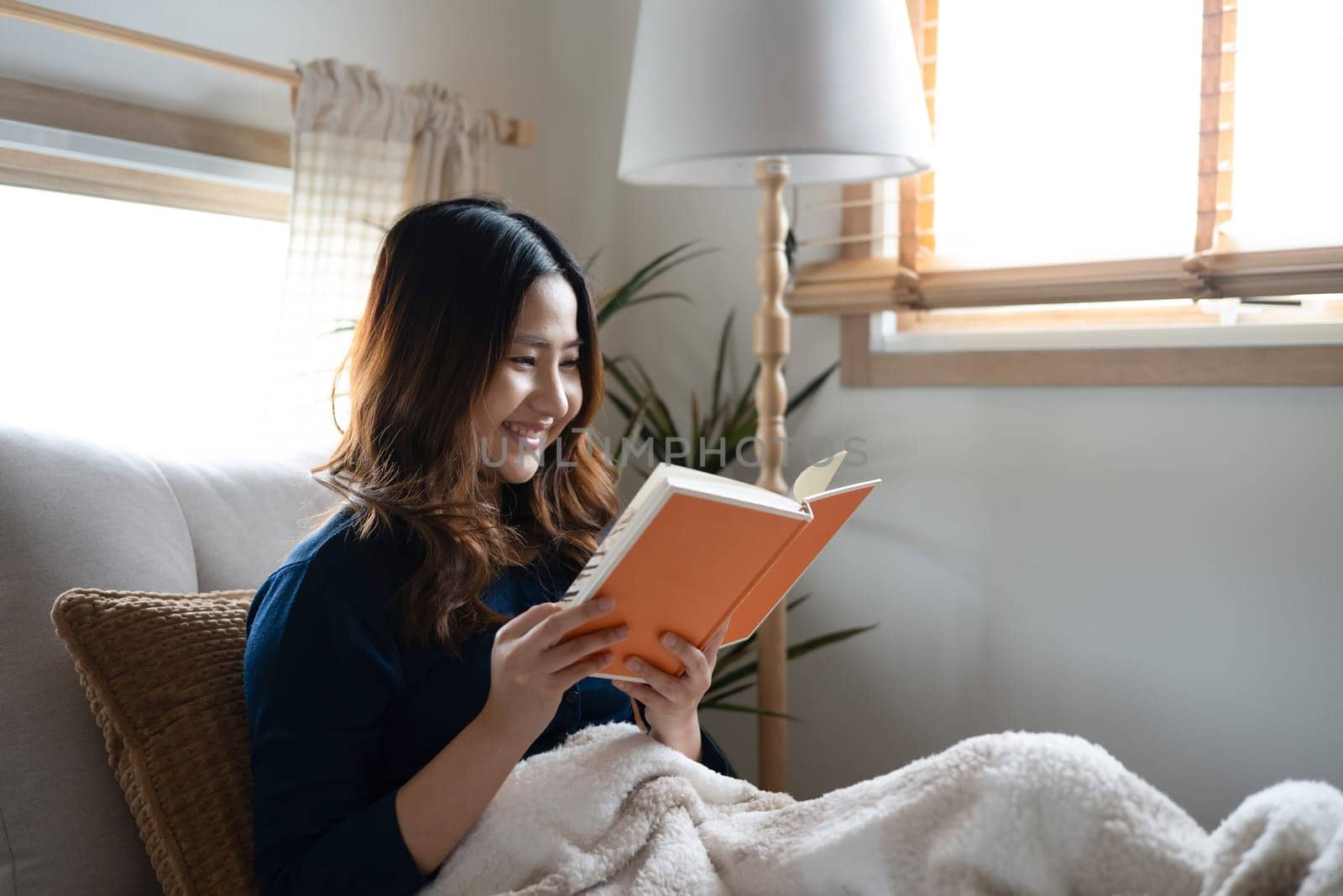 Smiling young asian woman wrapped in a soft blanket and reading a book on a couch.