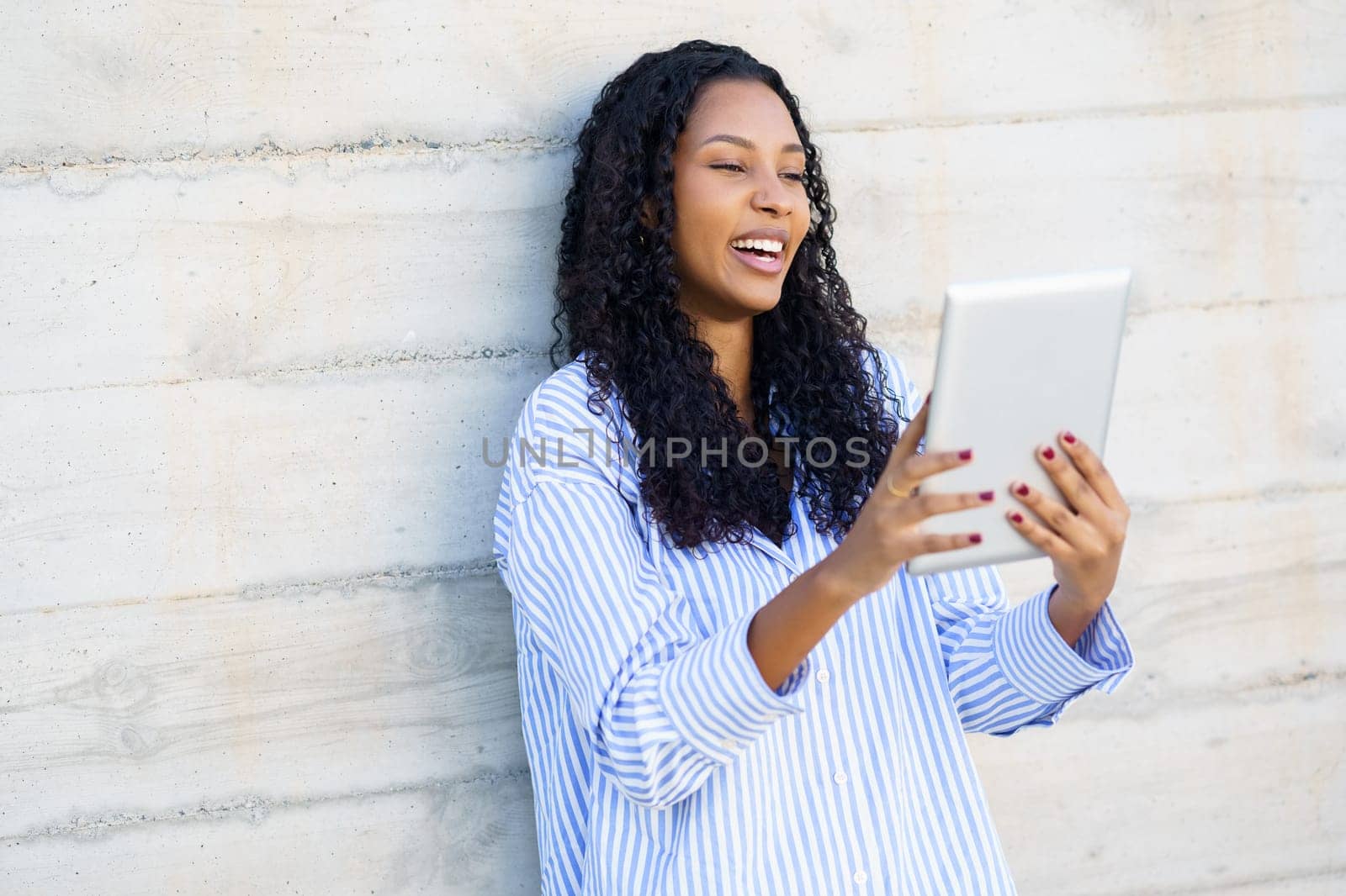 A happy woman smiles brightly while using her tablet against a modern wall, embodying the joy of technology and communication in enhancing her contemporary interactions and connectivity