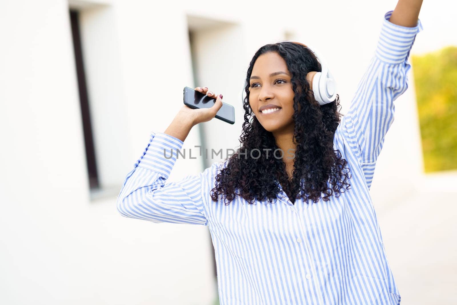 A joyful young woman dances energetically in the warm sunlight, wearing trendy headphones and skillfully holding her smartphone, truly embodying the joy of music and celebrating life in the moment