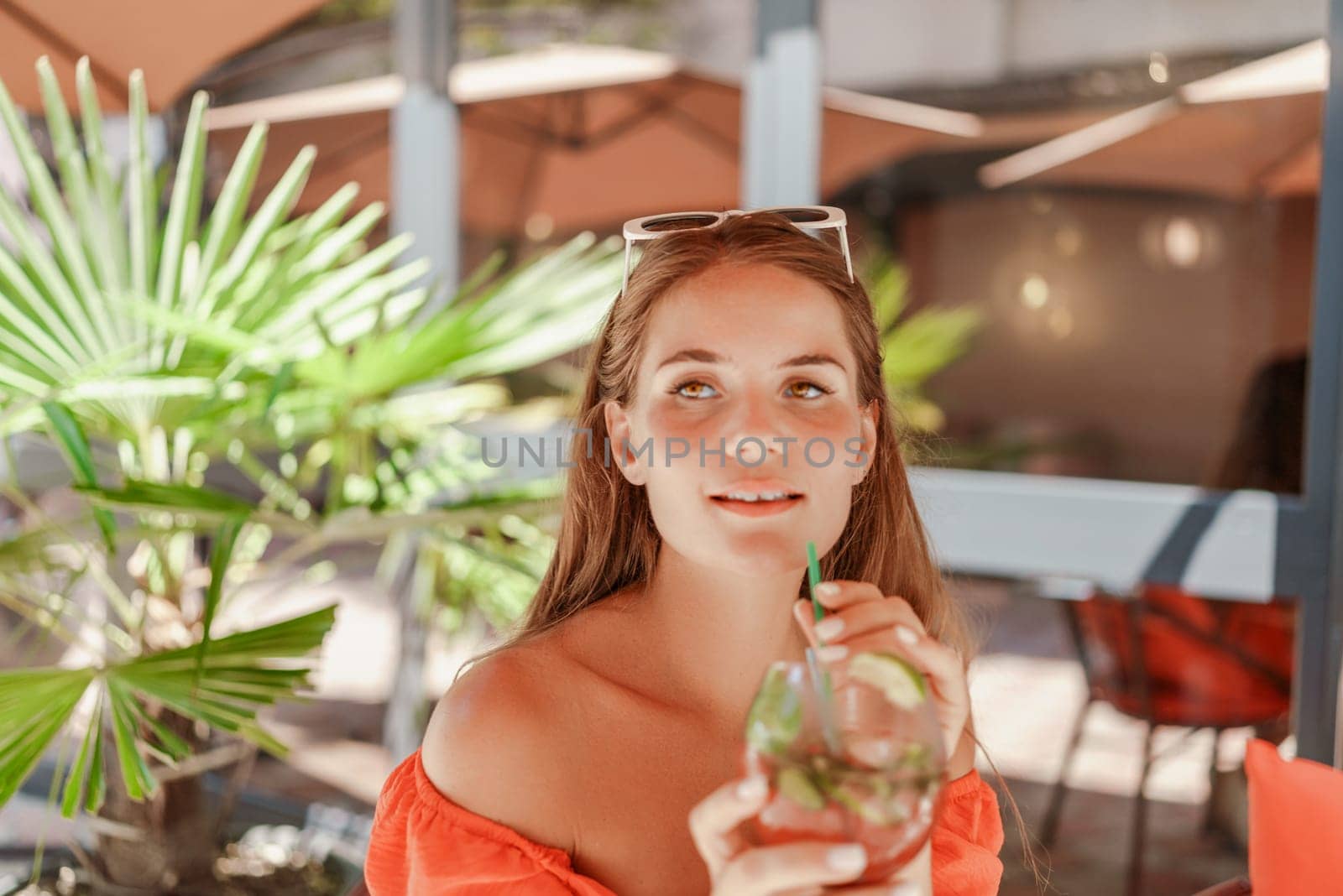 A woman in an orange dress is sitting in a chair with a drink in front of her. She is looking at the camera and she is enjoying her drink. The scene is set in a patio or outdoor area