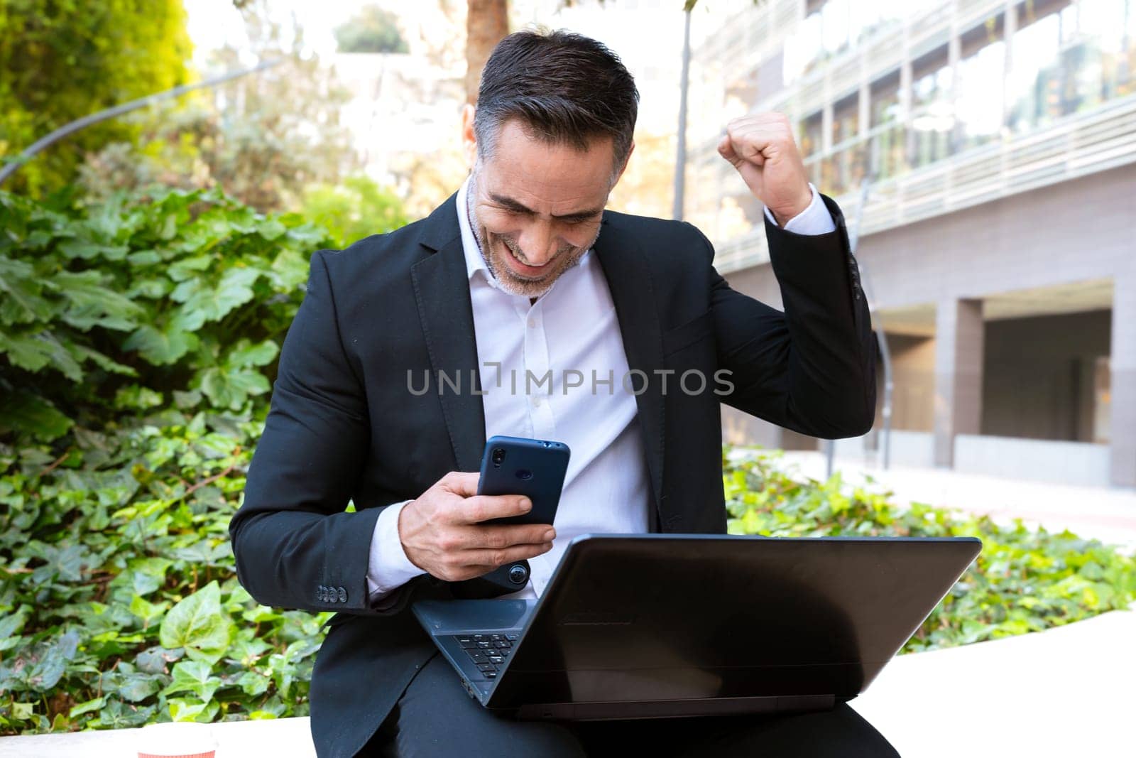 Excited businessman in a suit sitting outdoors, holding a smartphone and raising his fist in celebration. Represents success, achievement, and happiness in professional and digital work.