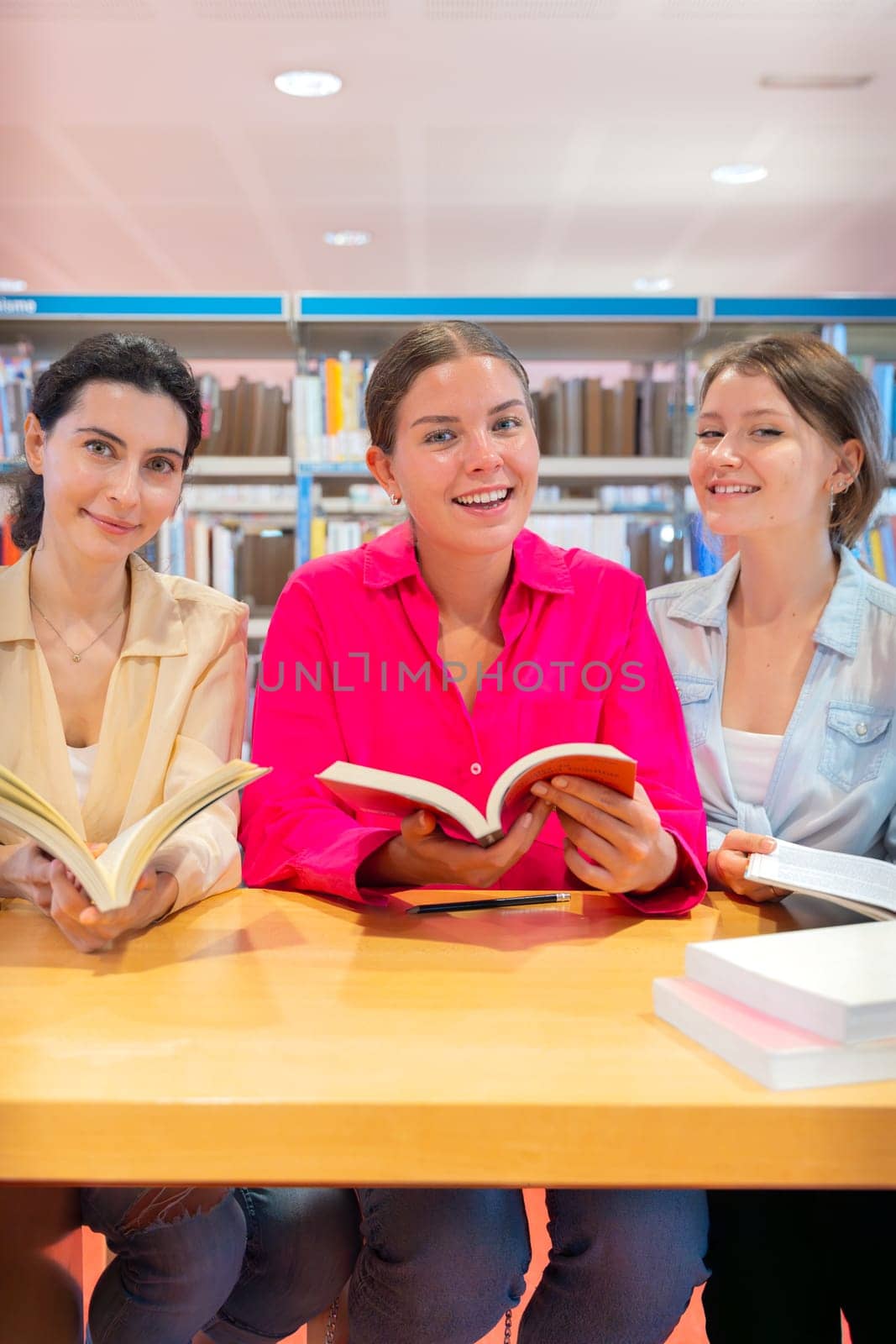 Three young women enjoying reading and learning at library. They are holding books and smiling. The atmosphere is cozy, and shelves filled with books can be seen in the background.Vertical