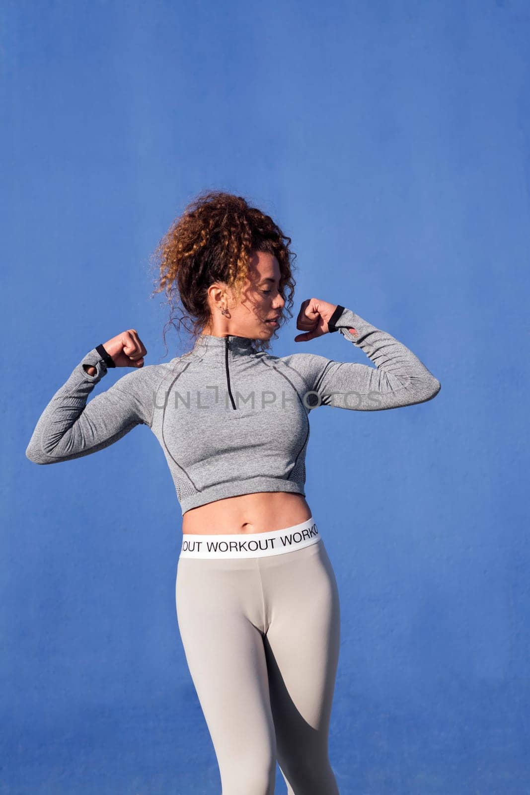 strong female athlete in sportswear with curly hair demonstrating muscles during workout in in front of blue background, concept of sport and healthy lifestyle