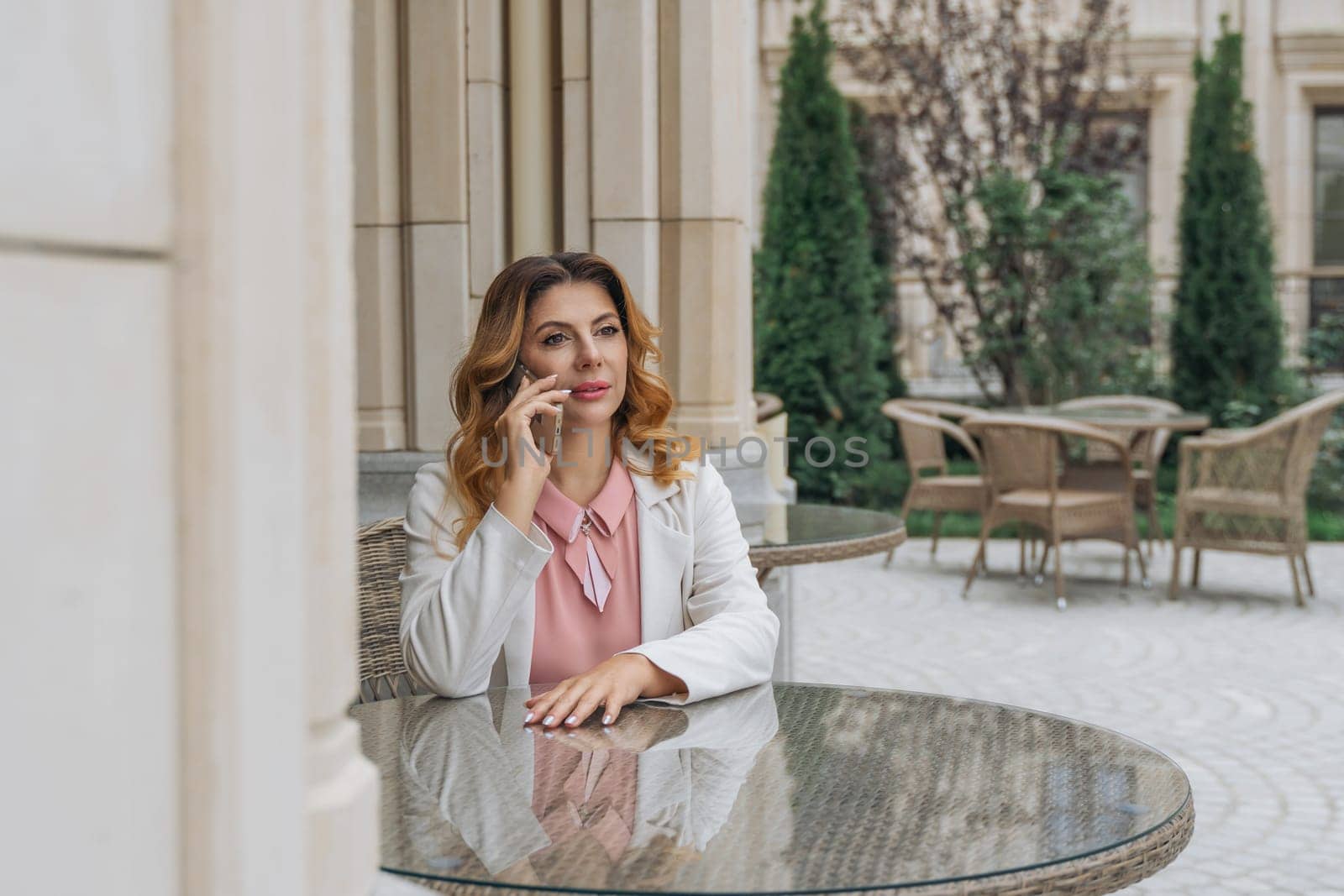 A woman with reddish-brown hair sits at a table with a glass top, talking on a cell phone in an outdoor cafe.