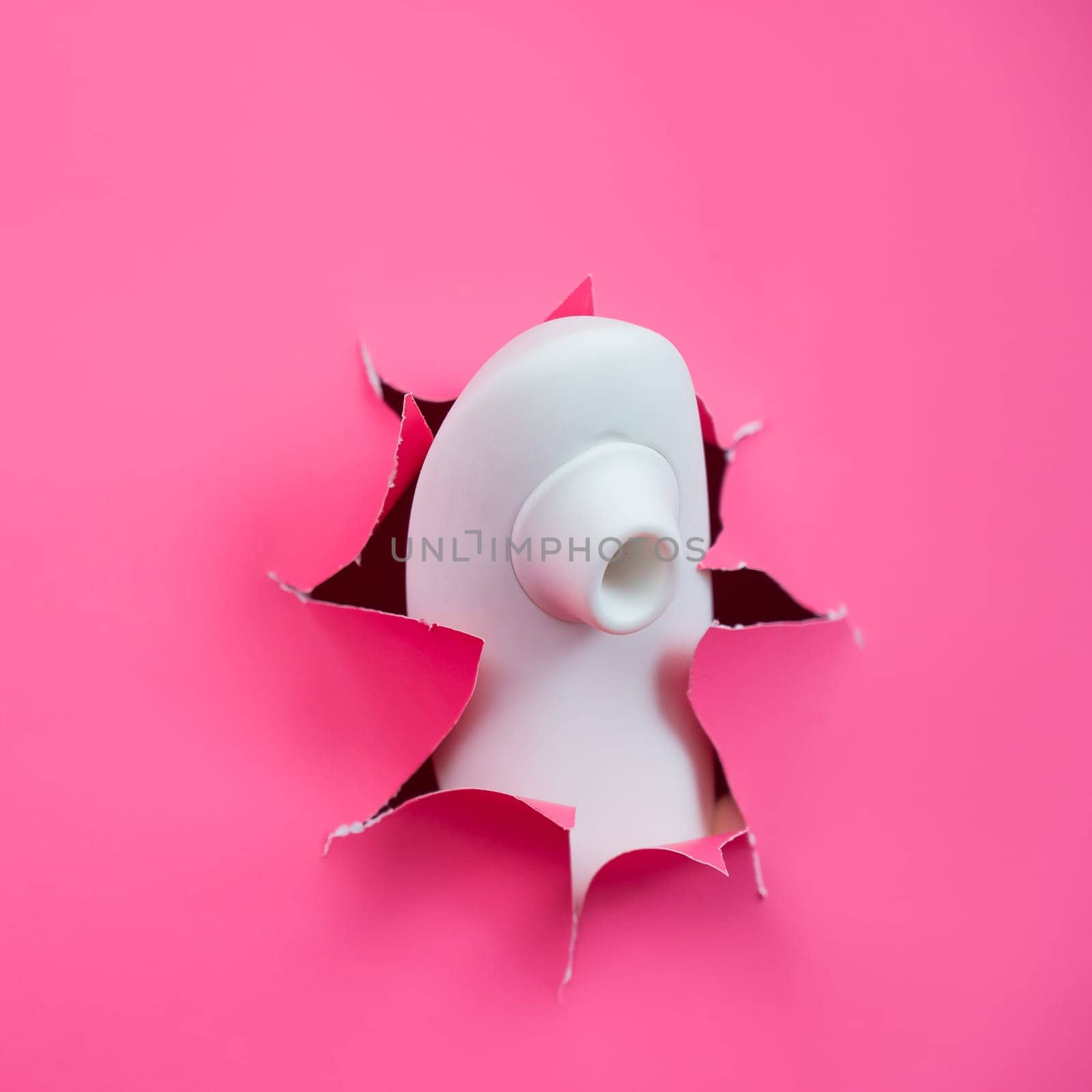 White vacuum clitoral vibrator sticking out through pink paper background. Sex toy