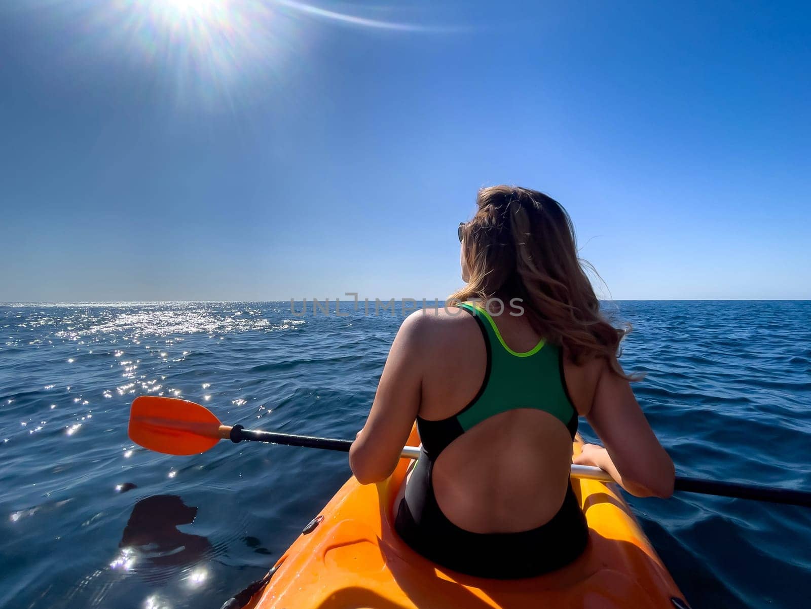 A woman paddles a yellow kayak on a calm ocean. She is wearing a green and black swimsuit and the sun is shining brightly overhead.