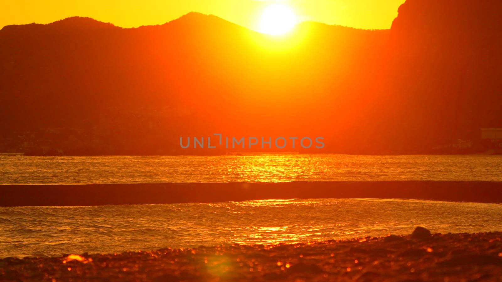 A fiery sunset casts an orange glow over a calm body of water, reflecting off the surface. Silhouettes of mountains rise in the distance.