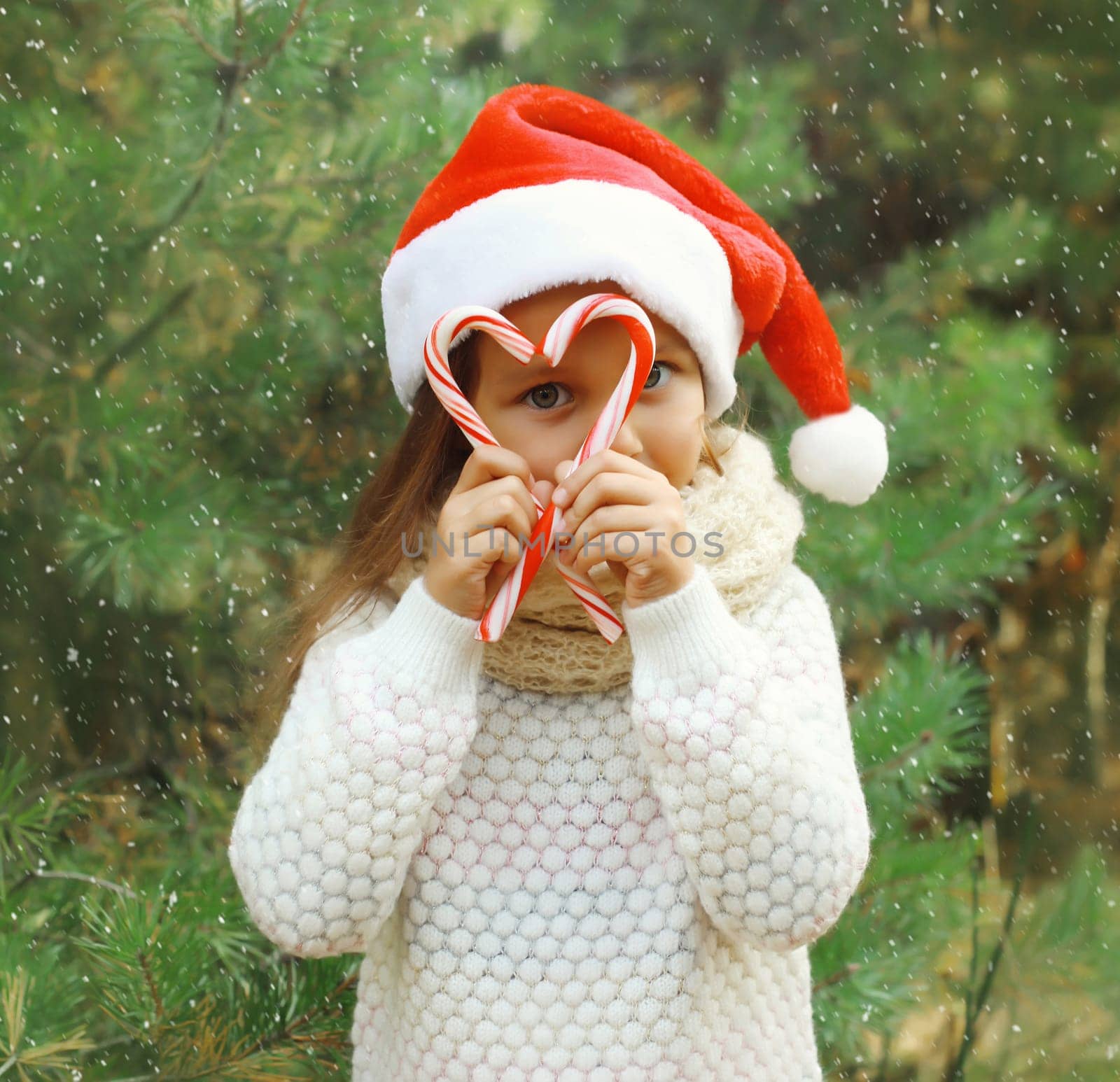 Christmas portrait of happy smiling little girl child in santa red hat with sweet lollipop cane stick outdoors on green tree background with snow