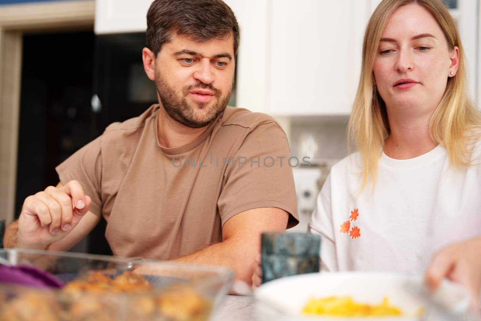 A man and a woman are seated at a kitchen table. The man is wearing a brown t-shirt and the woman is wearing a white t-shirt.