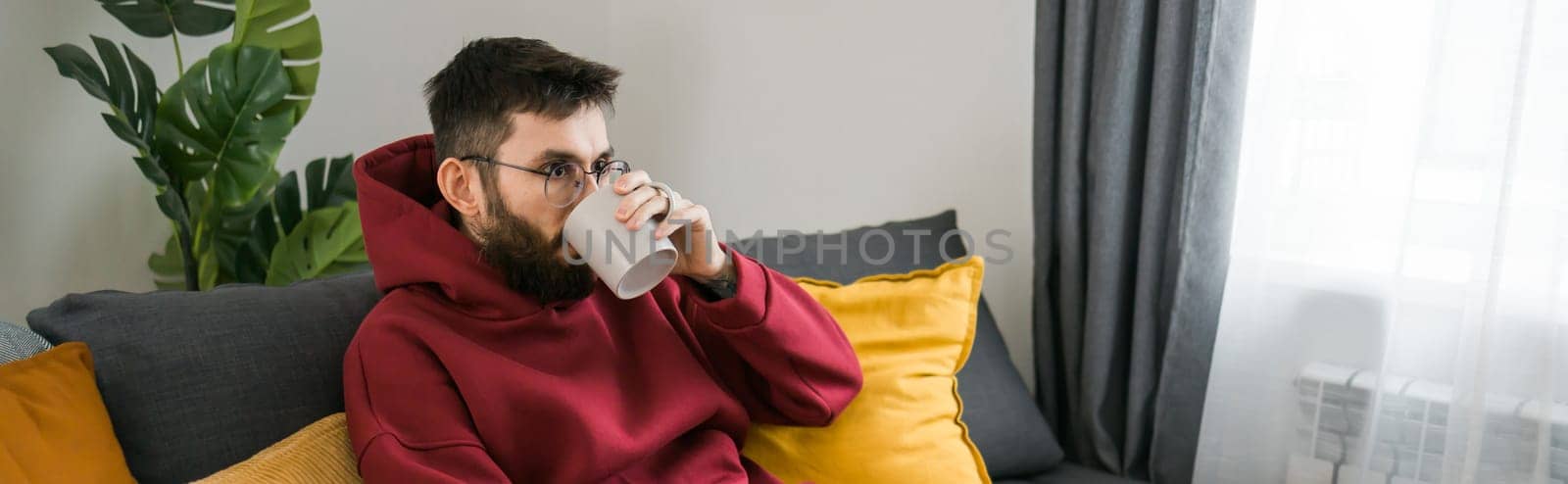 Young arab man drinking tea sitting on sofa at home copy space banner.