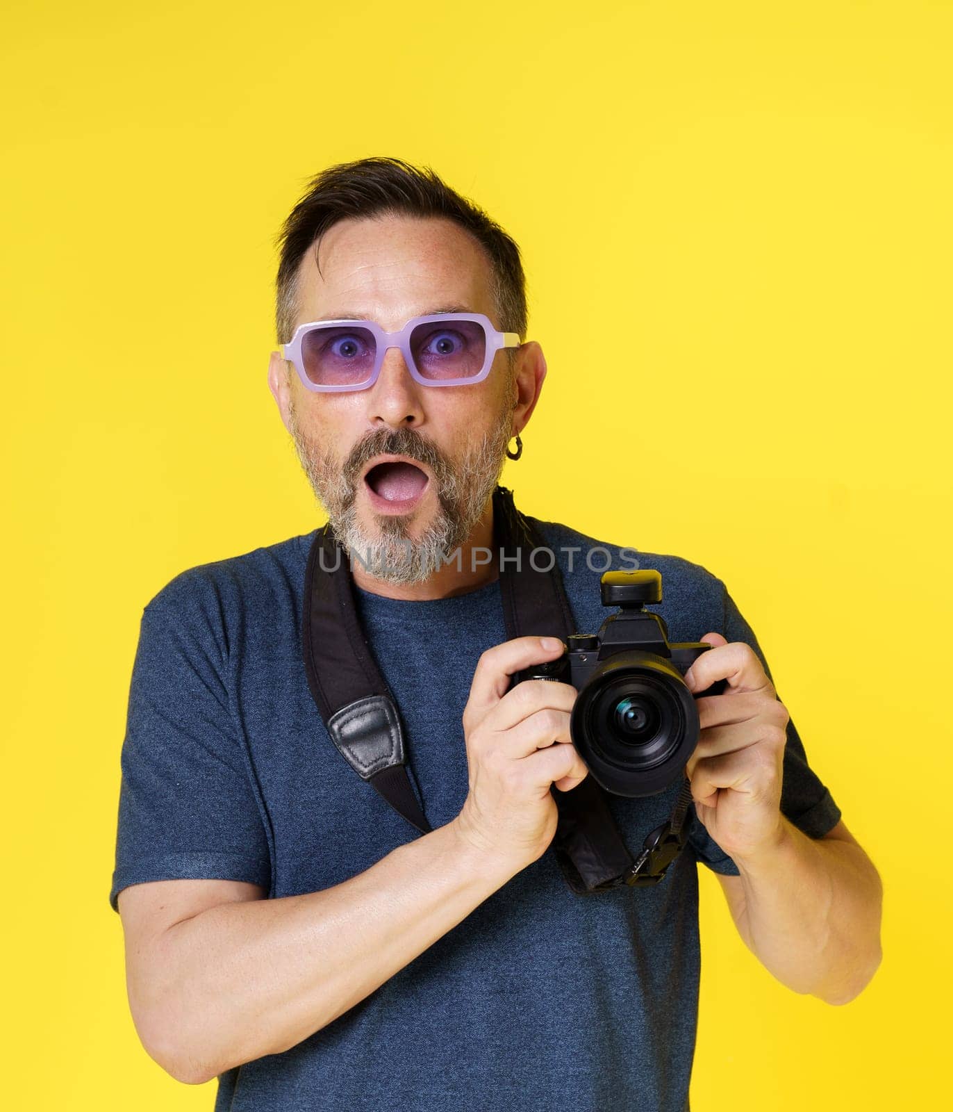 Passionate old photographer beams with excitement, holding mirrorless photo camera, in isolated studio shot on yellow background. Seasoned photographer continues pursue photography passion with zeal. by LipikStockMedia