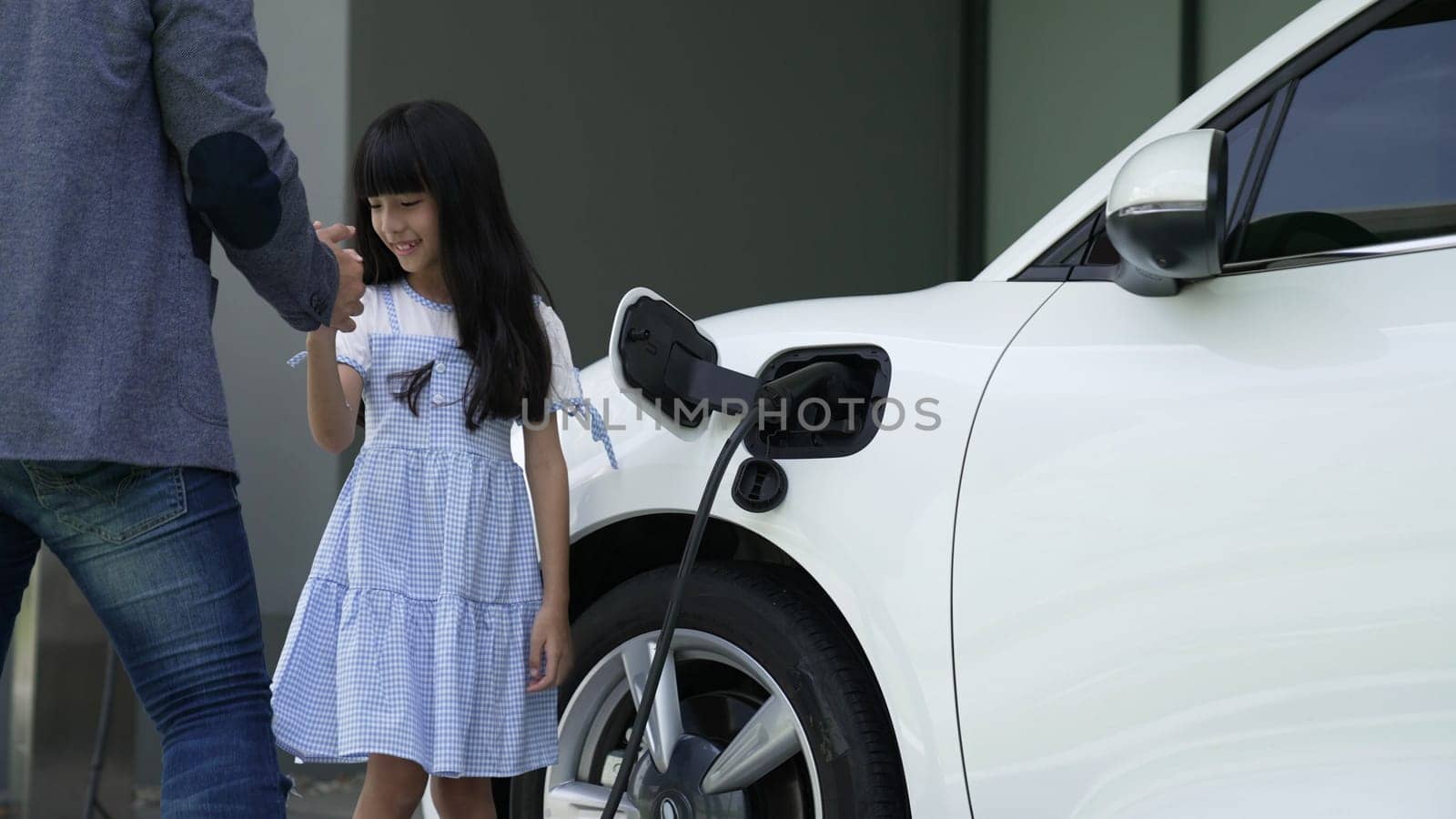 Progressive father and daughter installing plug from home charging station for electric vehicle. Future eco-friendly car with EV cars powered by renewable source of clean energy.