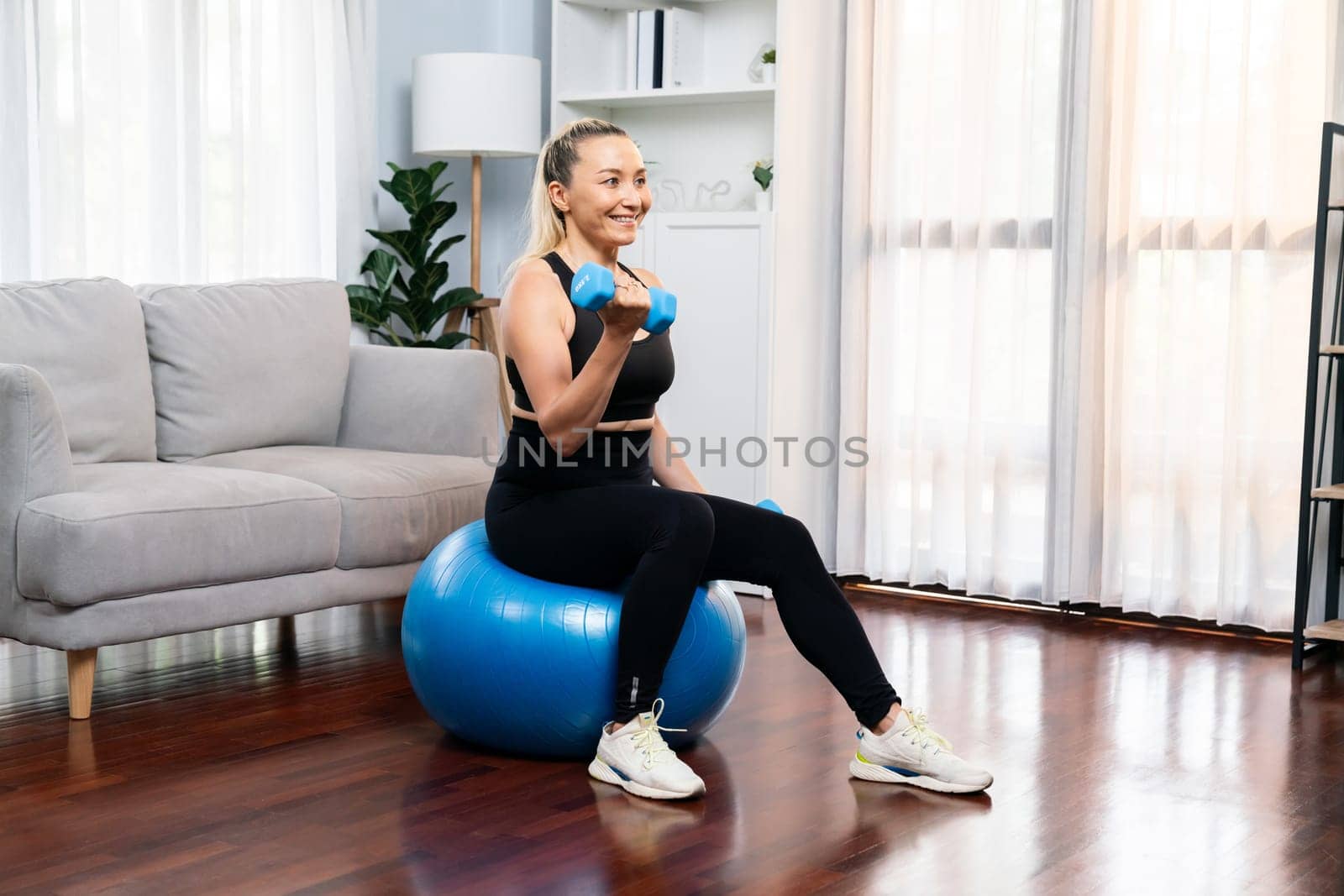 Athletic and sporty senior woman sitting on fit ball while engaging in weight lifting with dumbbell at home exercise as concept of healthy fit body lifestyle after retirement. Clout