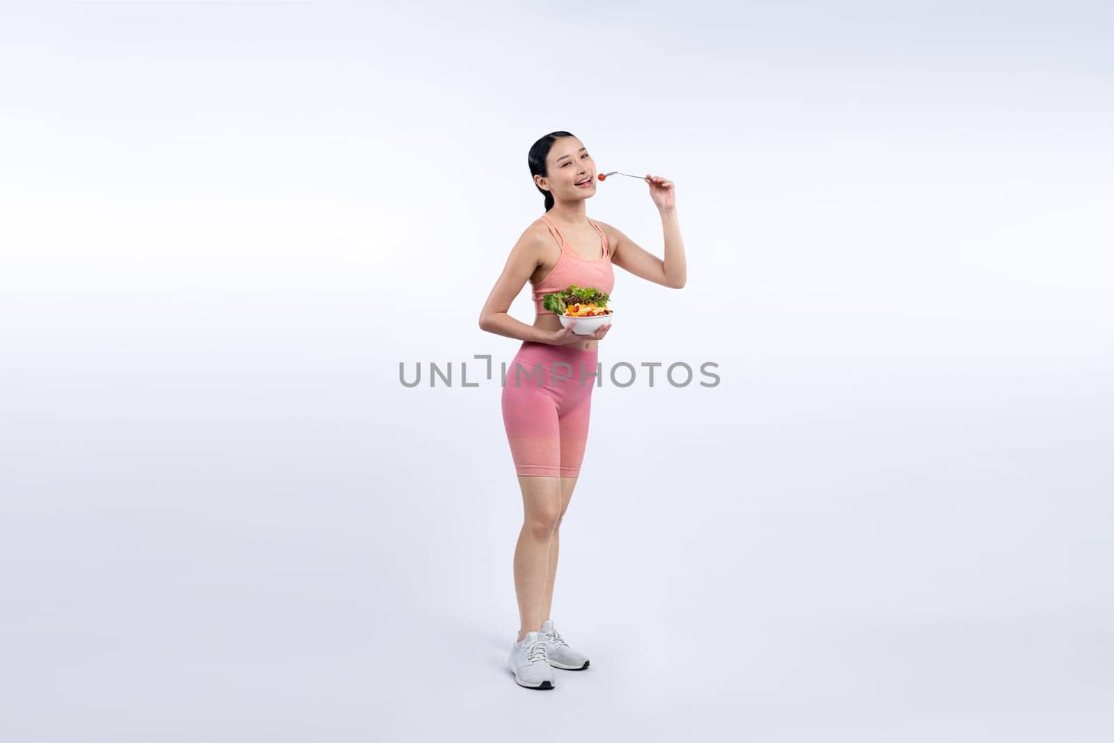 Young sporty Asian woman in sportswear holding salad bowl fill with vibrant of fruit and vegetable. Natural youthful and fit body lifestyle with balance nutrition on isolated background. Vigorous