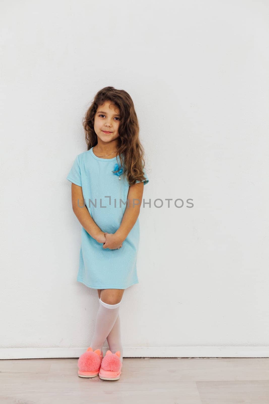 Little Curly Girl In Dress And Pink Slippers Against White Wall