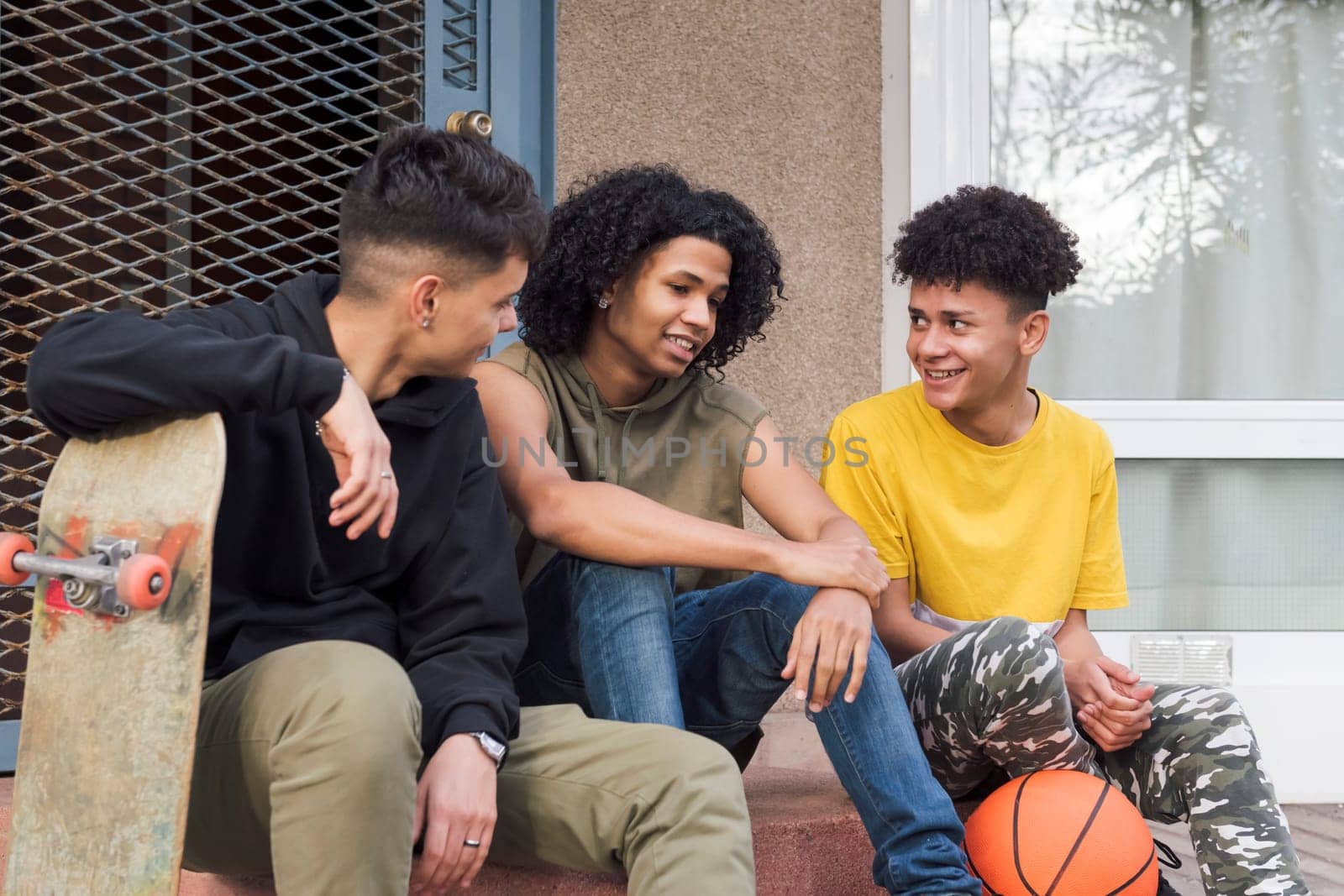 three multiracial male teenagers with basketball and skateboard having fun and talking sitting together on street, concept of youth and urban lifestyle