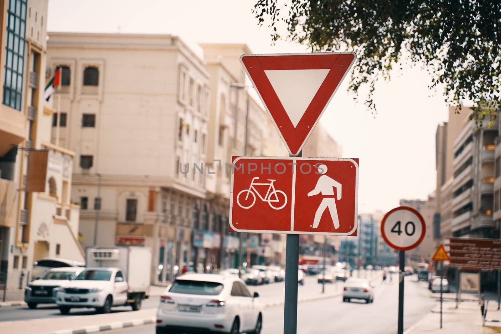 A bustling city street with a bicycle and pedestrian sign, surrounded by buildings and trees. Vehicles and pedestrians share the road in this urban setting