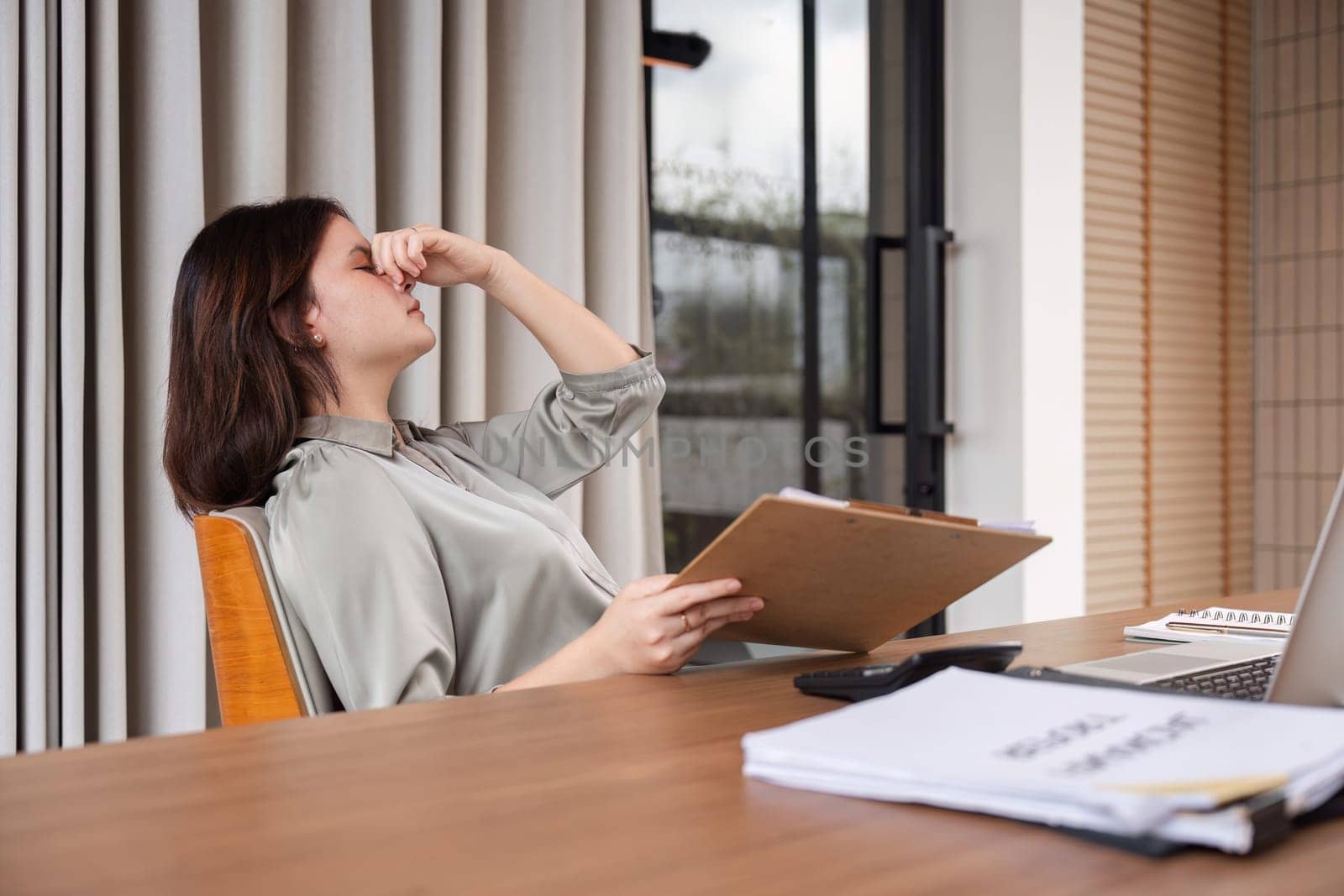 A businesswoman feeling stressed at her desk, surrounded by documents and a laptop in a contemporary office environment.