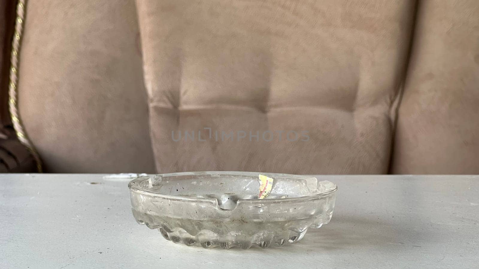 A clear glass ashtray rests on a table near a soft, beige couch, reflecting light.