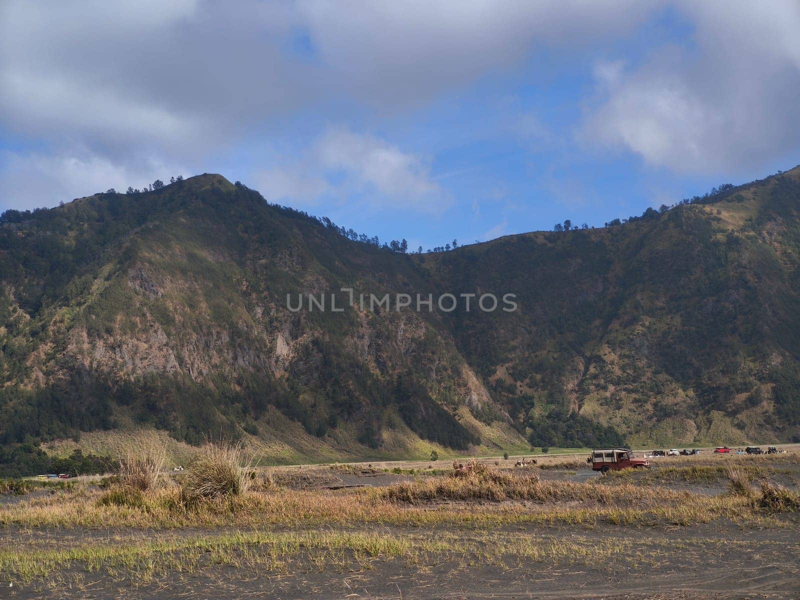 A majestic view of the Bromo Tengger Semeru National Park. The iconic Mount Bromo stands tall with its distinctive crater, surrounded by a vast expanse of volcanic sand and a clear blue sky dotted with fluffy clouds.