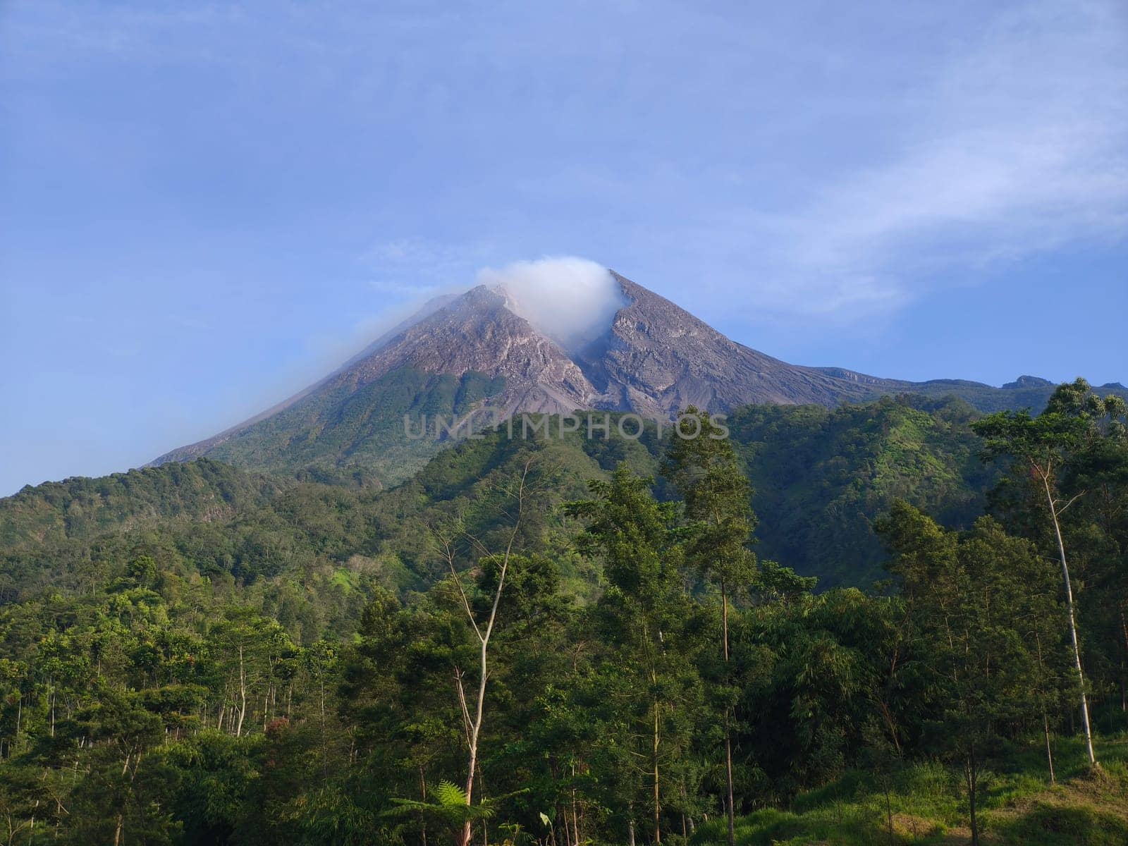 the majestic Mount Merapi, an active volcano in Indonesia. Its peak is shrouded in a wisp of cloud, and the slopes are covered in lush greenery.