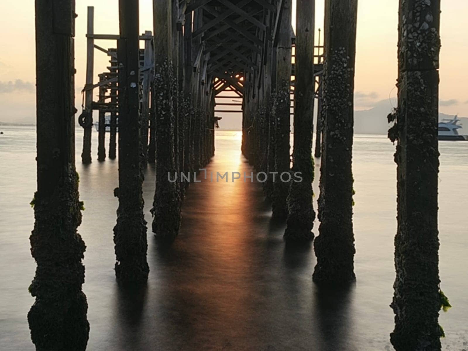 A weathered wooden pier extends into the water at sunrise, with barnacles clinging to the support posts near Banyuwangi, Indonesia
