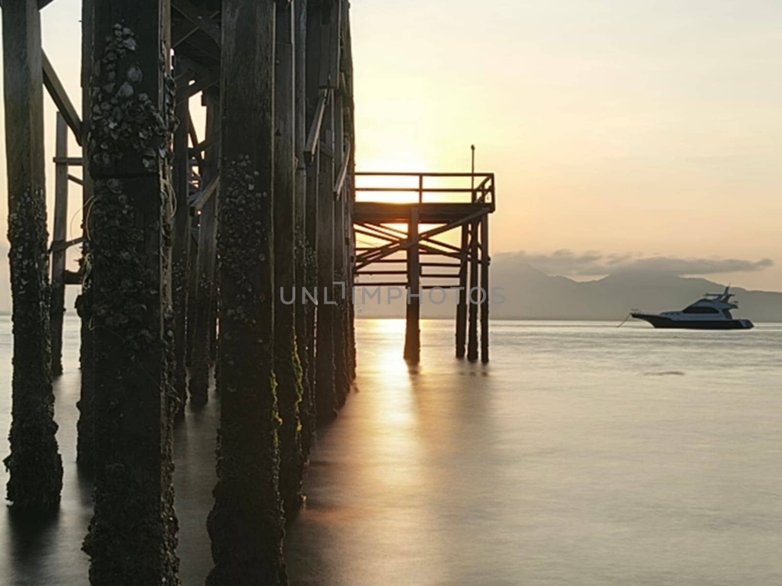A weathered wooden pier extends into the water at sunrise, with barnacles clinging to the support posts near Banyuwangi, Indonesia
