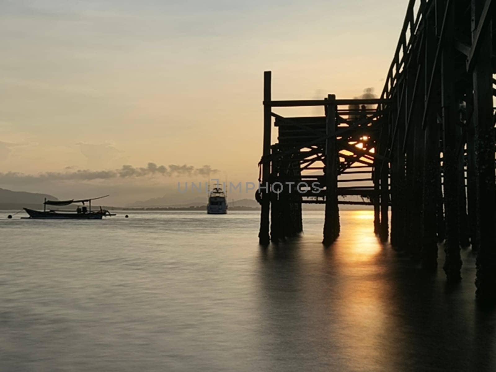 A weathered wooden pier extends into the water at sunrise, with barnacles clinging to the support posts near Banyuwangi, Indonesia