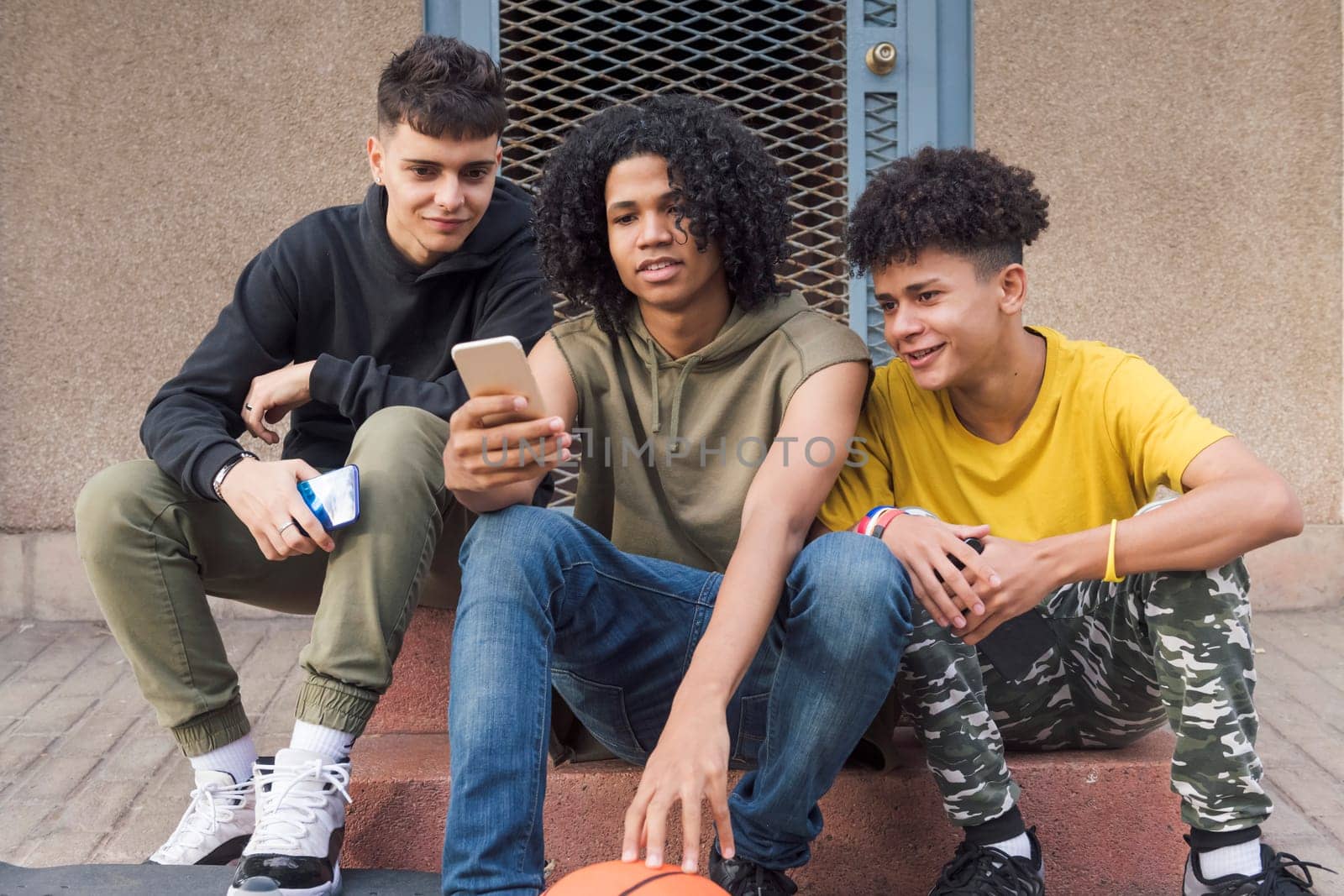 three happy multiethnic teen friends sharing mobile phone sitting together with on street, concept of youth and technology of communication