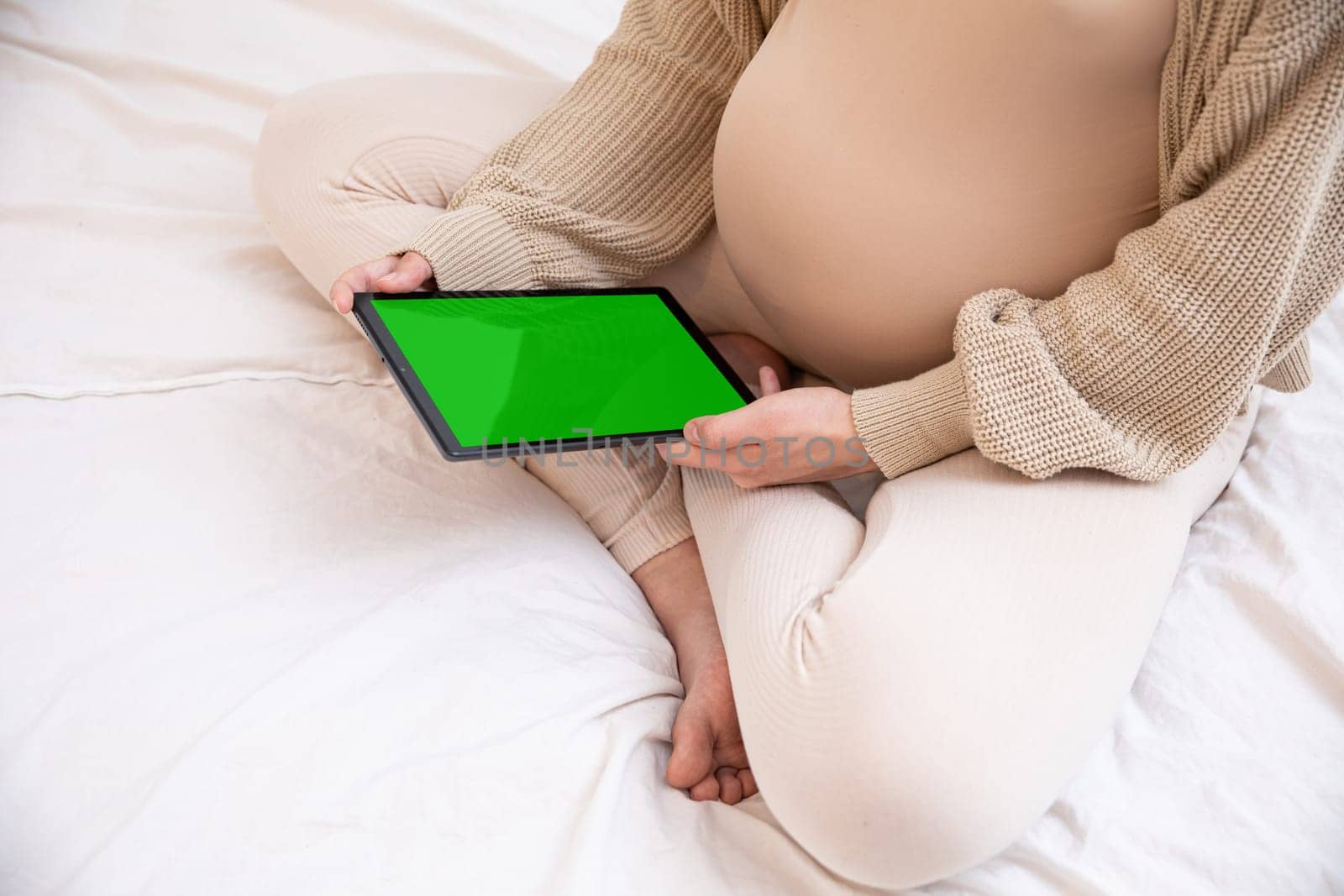 A pregnant woman is seated comfortably on her bed, gently holding a tablet that displays a vibrant green screen