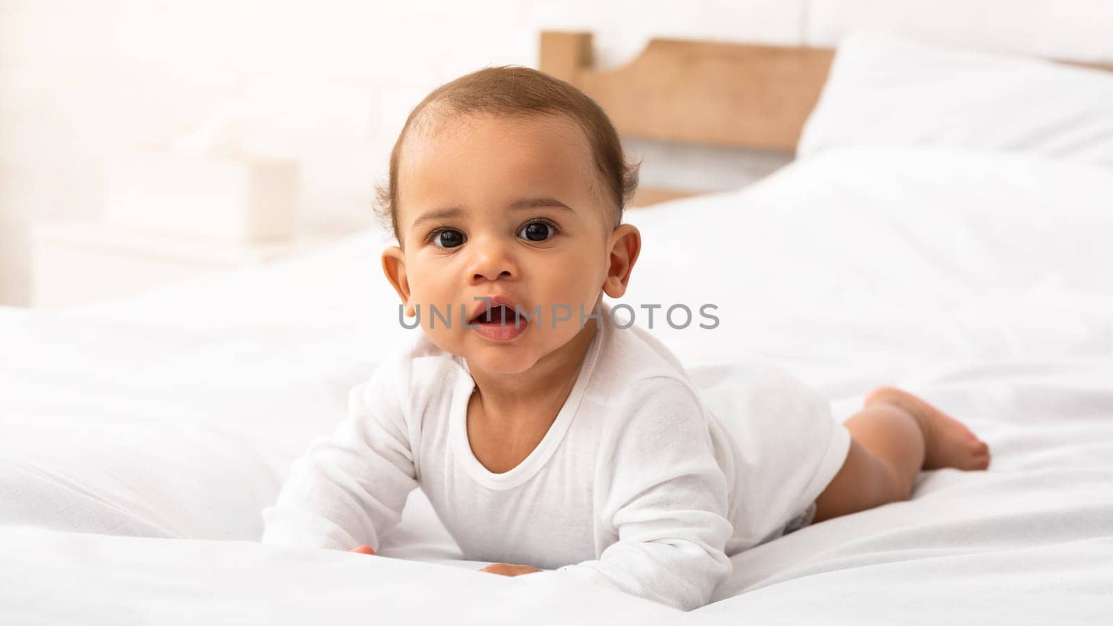 Cute African American Baby Infant In White Longsleeve Coverall Lying On Stomach In Bed Looking At Camera. Sweet Toddler Boy Posing Crawling In Bedroom At Home. Child Care, Clothes For Babies