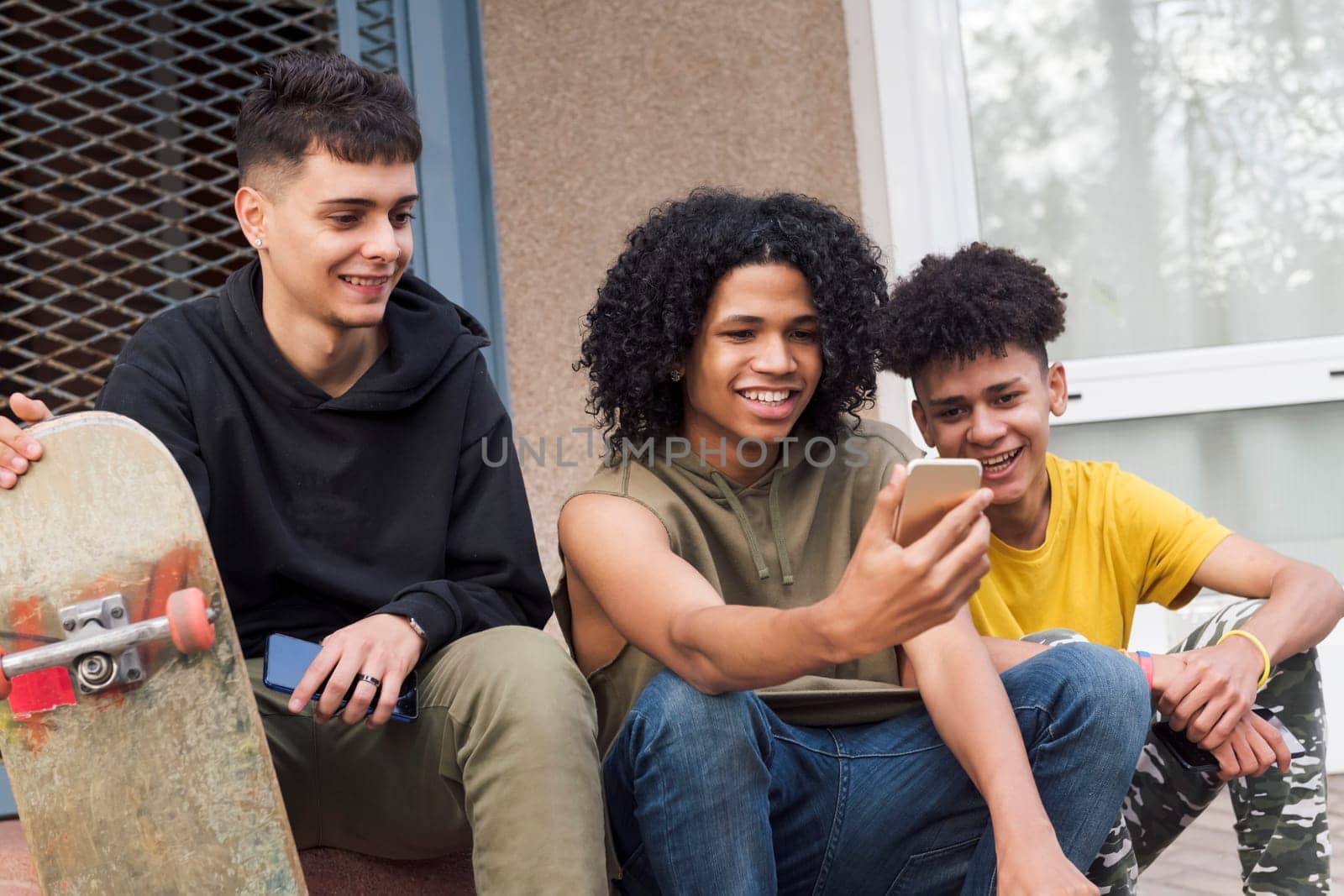 three happy multiethnic teen friends sharing mobile phone sitting together with on street, concept of youth and technology of communication