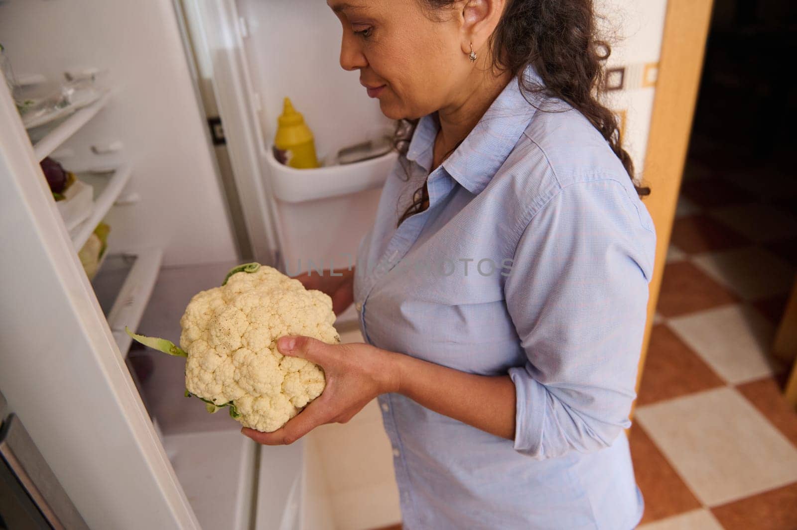 A woman takes out a fresh cauliflower from the refrigerator, symbolizing healthy living and nutrition. The kitchen setting evokes a sense of home cooking and well-being.