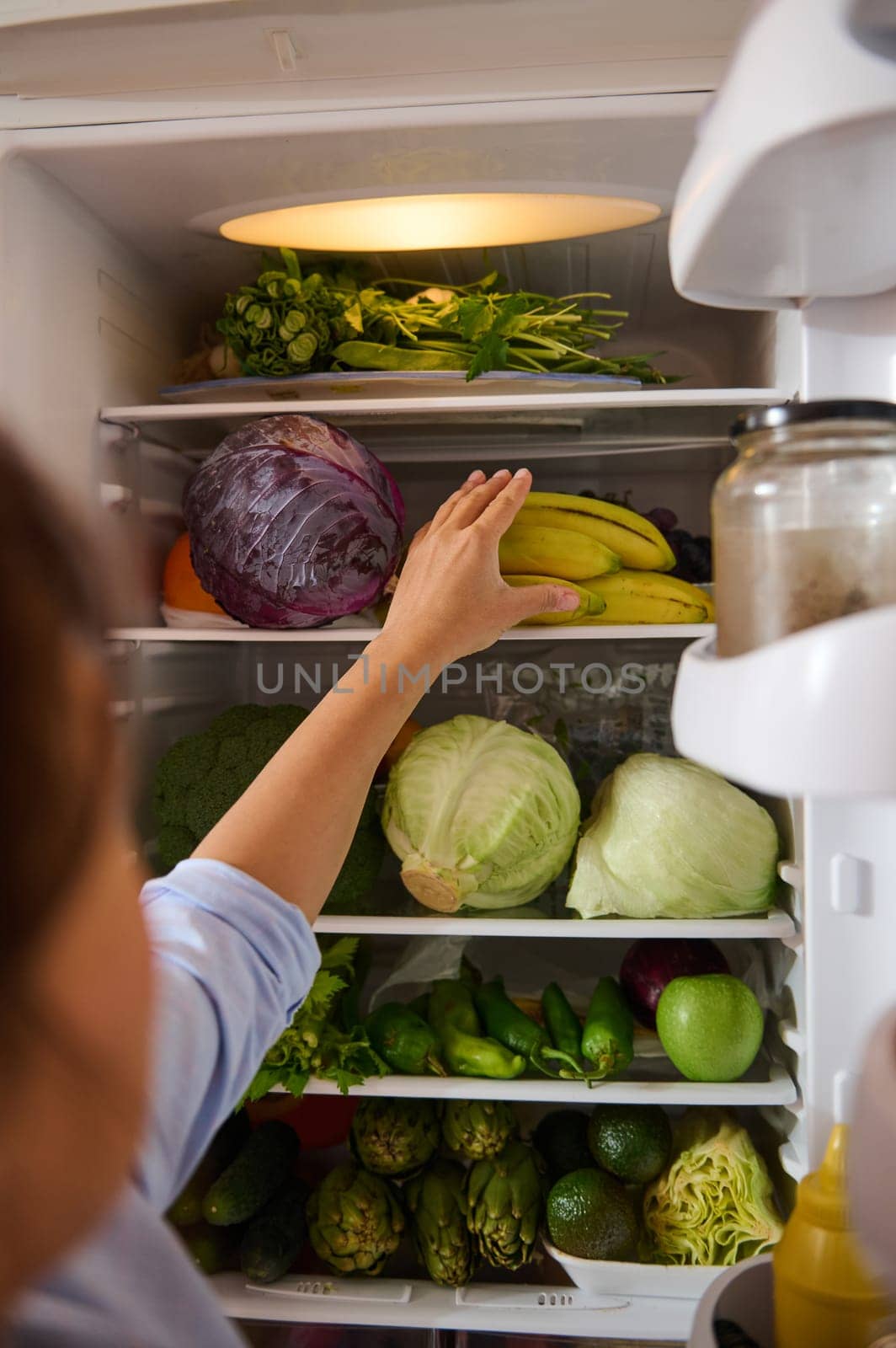 A person reaches into a refrigerator filled with fresh vegetables and fruits, including bananas, cabbage, and greens. The image conveys healthy eating, freshness, and home organization.