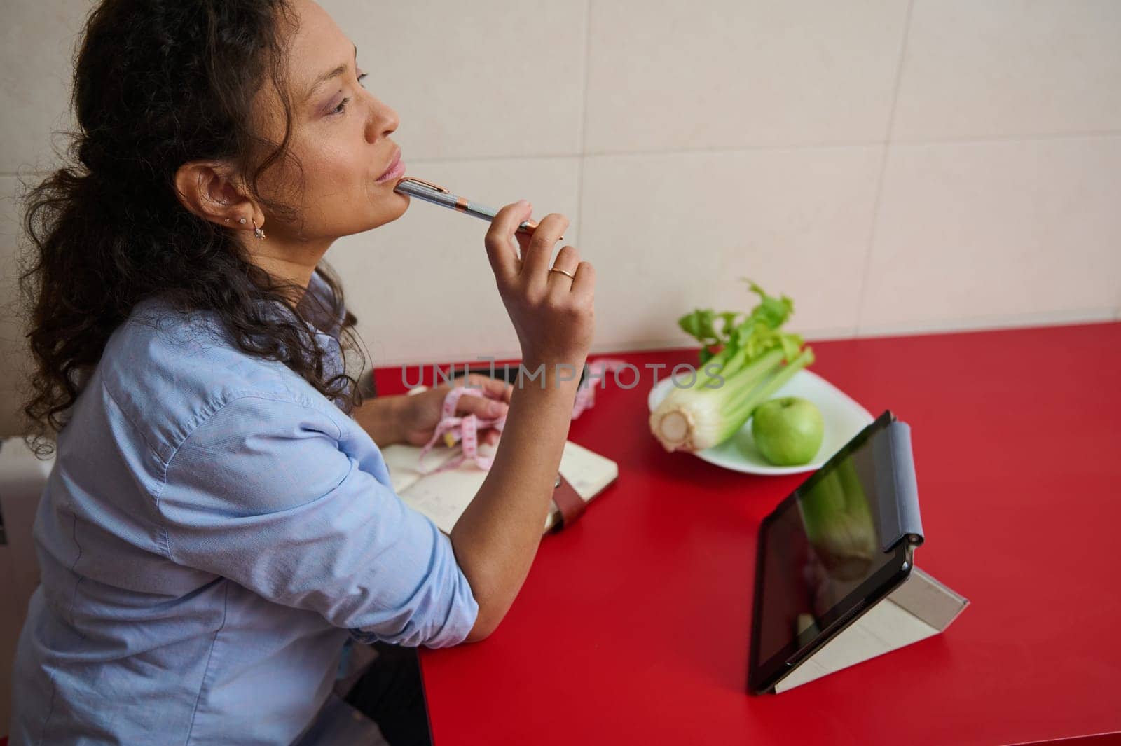 A woman sits at a red table with vegetables and a tablet, pondering healthy recipes. She uses a pen in thought while managing a notepad, evoking creativity and focus on nutrition.