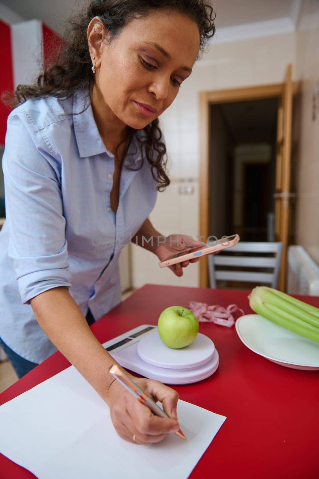 A woman in a kitchen uses her smartphone to track the calories of an apple on a digital scale. The scene emphasizes healthy living, nutrition, and modern technology in daily life.