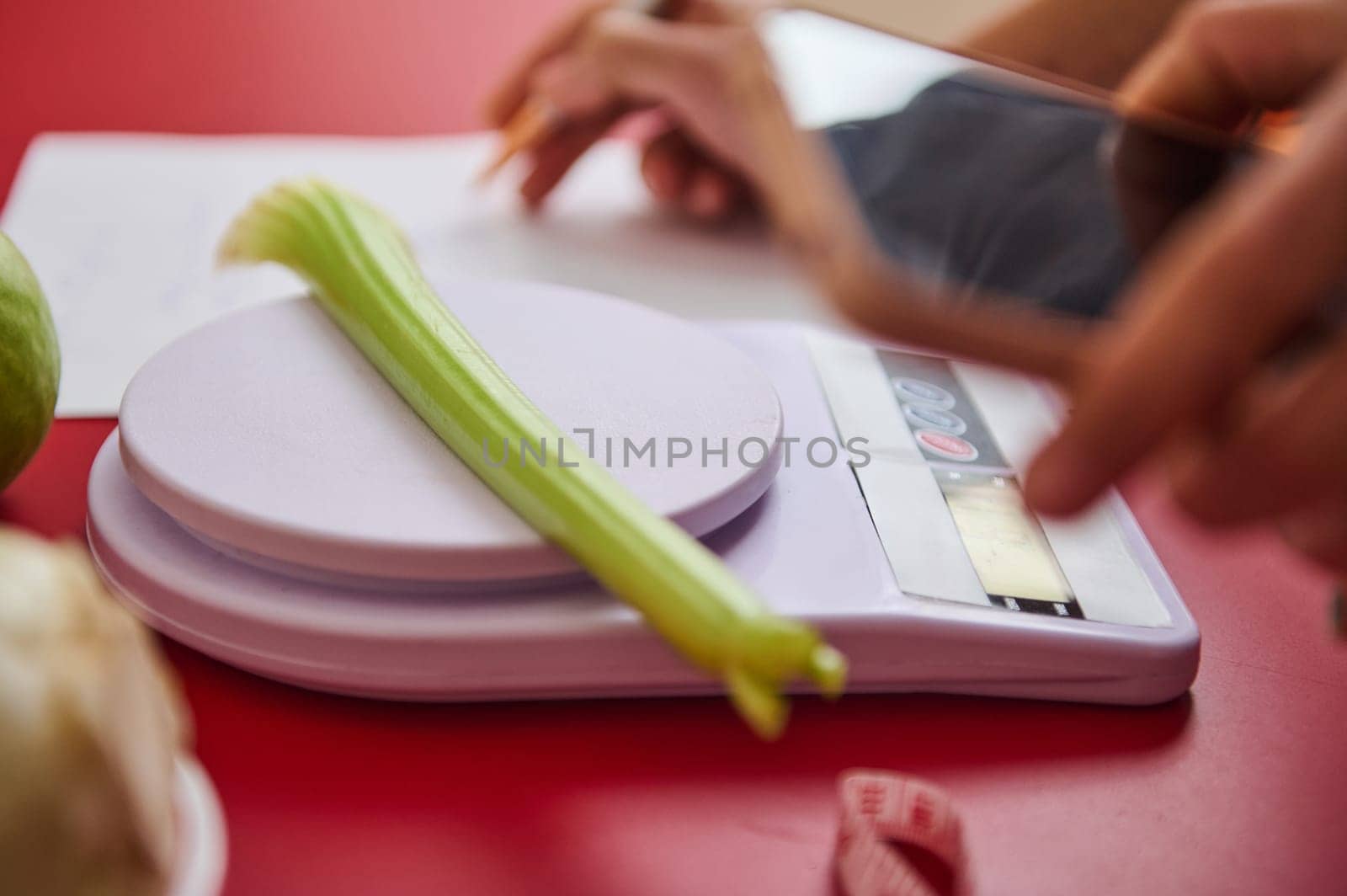 A fresh celery stalk rests on a digital scale as hands hold a smartphone. The image emphasizes healthy eating and meal planning with a focus on precision and nutrition.
