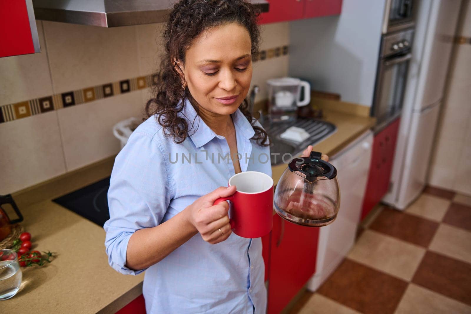 A woman stands in a modern kitchen, enjoying a fresh cup of coffee. She appears relaxed and content, taking a moment from her daily routine. Red cabinet and countertop add to the cozy ambiance.