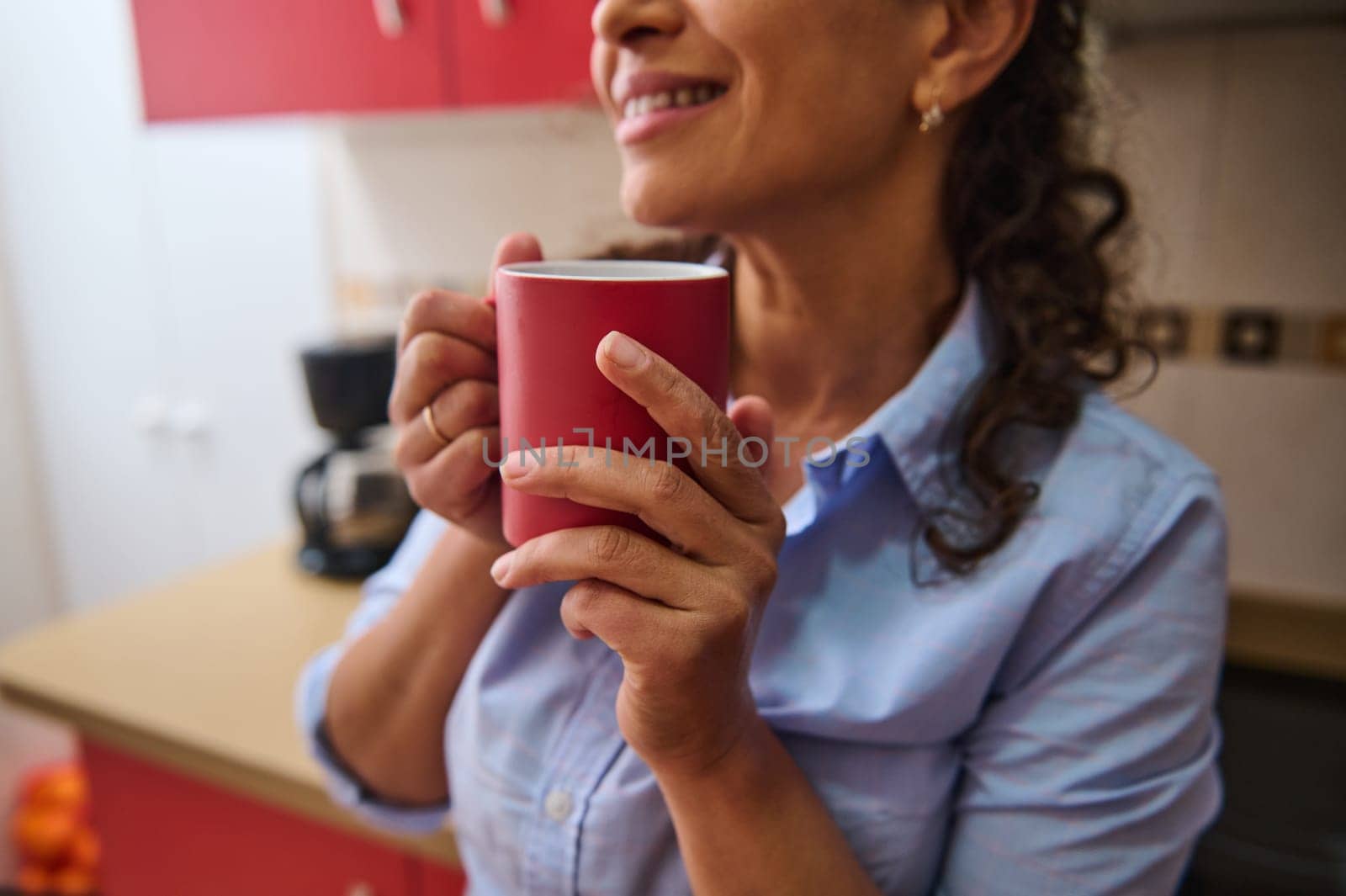 A woman holds a red mug, enjoying a warm drink in a cozy kitchen. Her relaxed expression suggests comfort and calm, creating a peaceful, homey atmosphere.