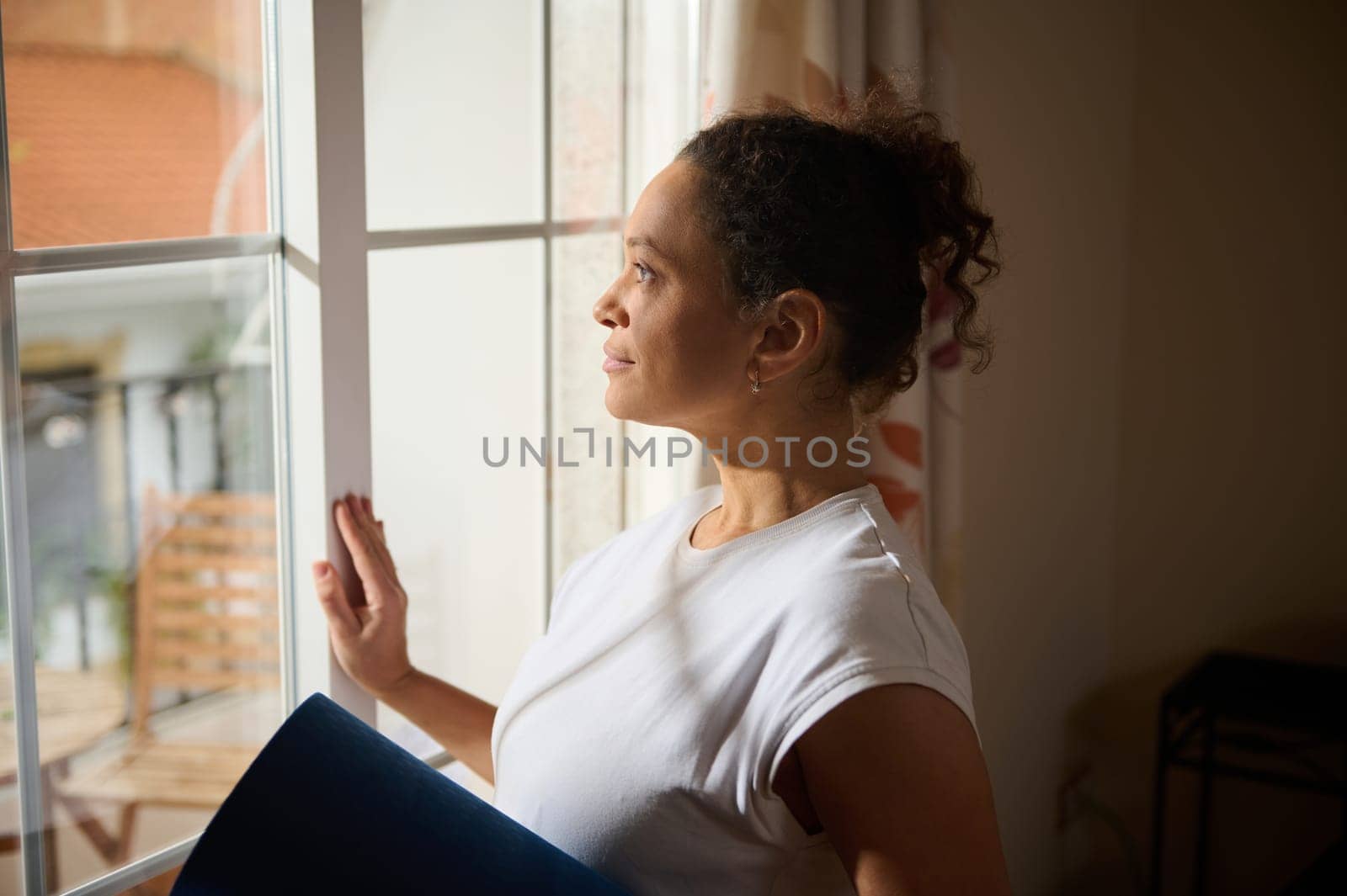 A woman stands by a window, holding a yoga mat and gazing outside. She appears contemplative and calm, enjoying a moment of reflection. The setting is warm and inviting, suggesting tranquility.