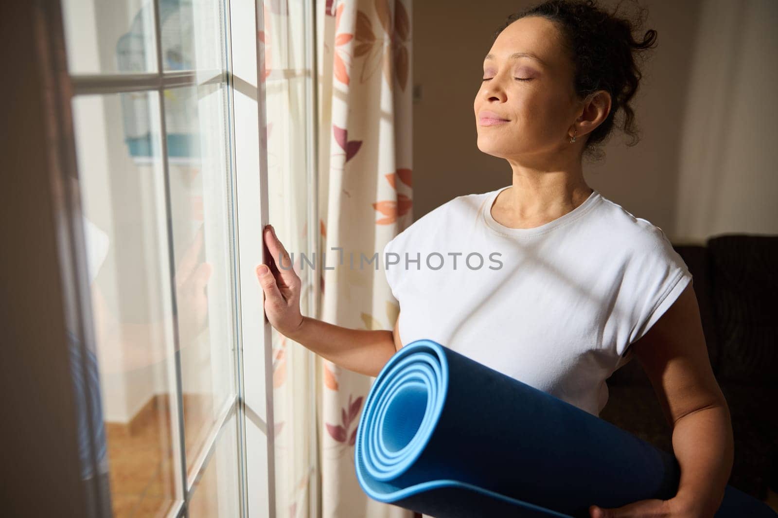 A serene woman stands by a window, eyes closed, holding a yoga mat. She basks in the sunlight, embodying peace and relaxation. It's a moment of mindfulness and tranquility at home.